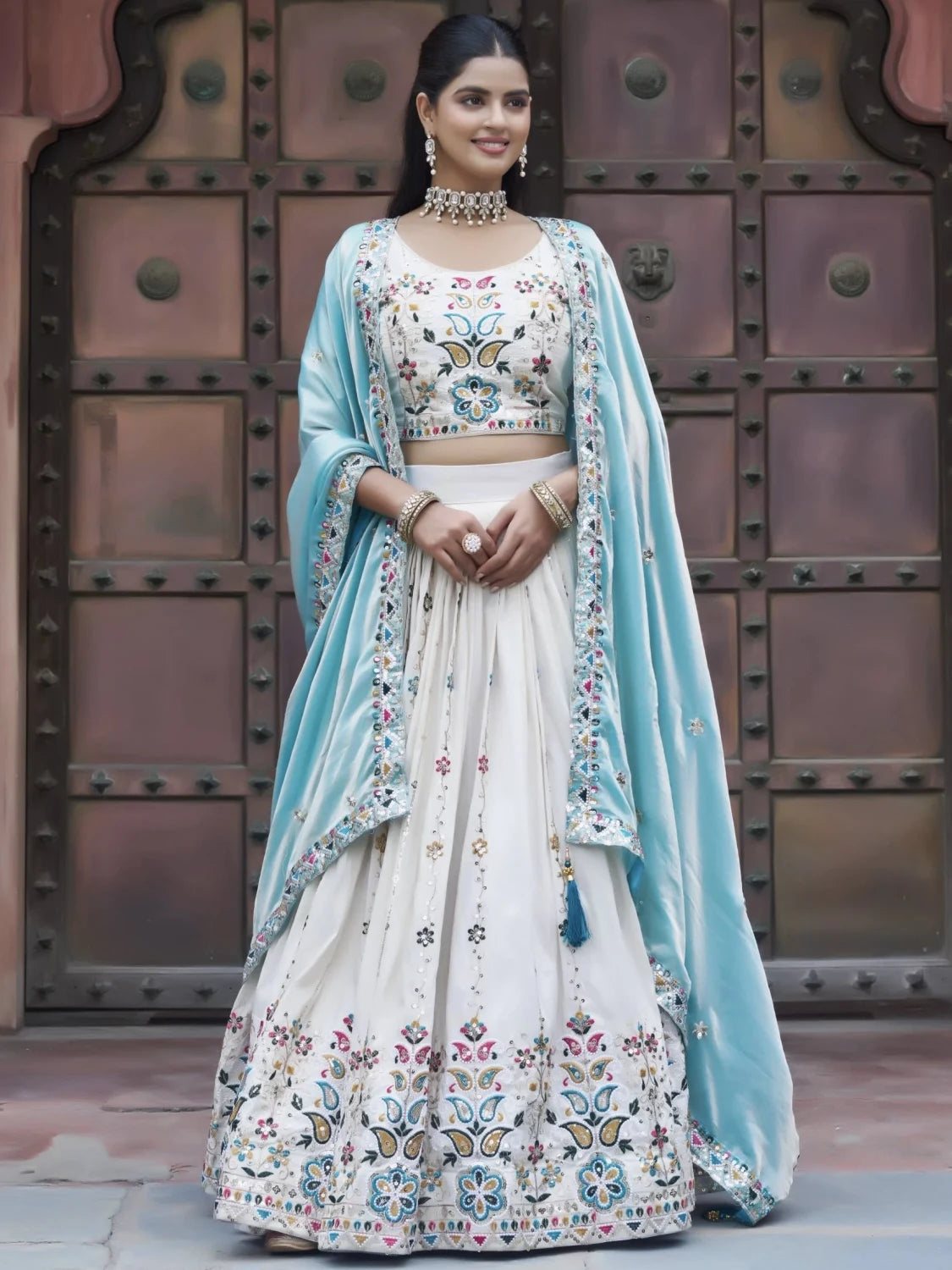 Woman wearing a traditional embroidered outfit with a light blue dupatta against a metallic door background