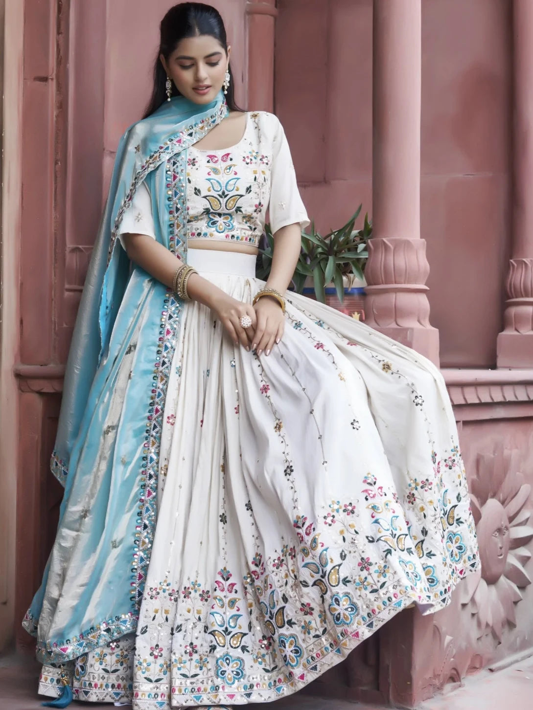 Woman in a traditional outfit with floral patterns standing against a pink architectural background