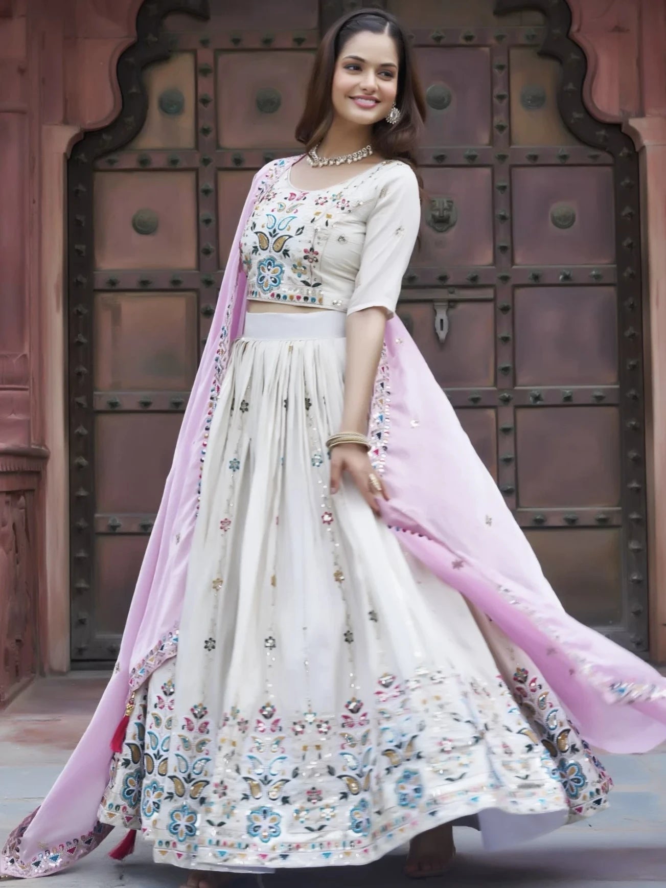 Woman in a traditional outfit with floral patterns and a pink dupatta against a textured wall.