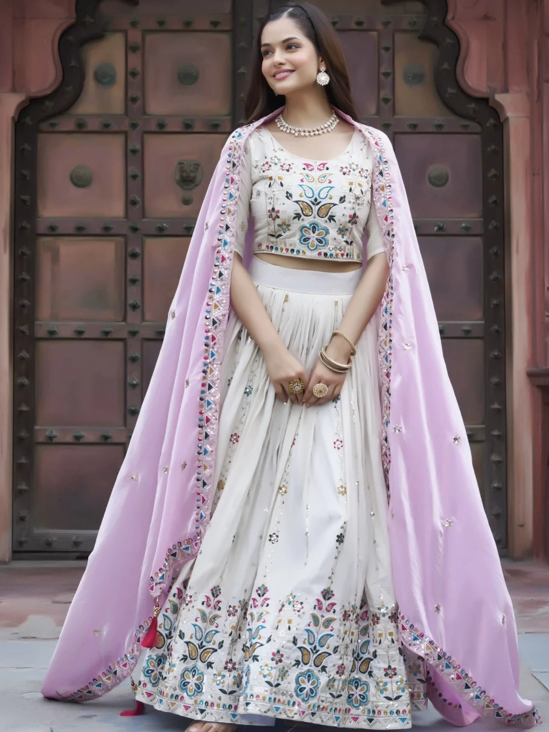Woman in traditional outfit with floral patterns and pink dupatta against a textured wall.
