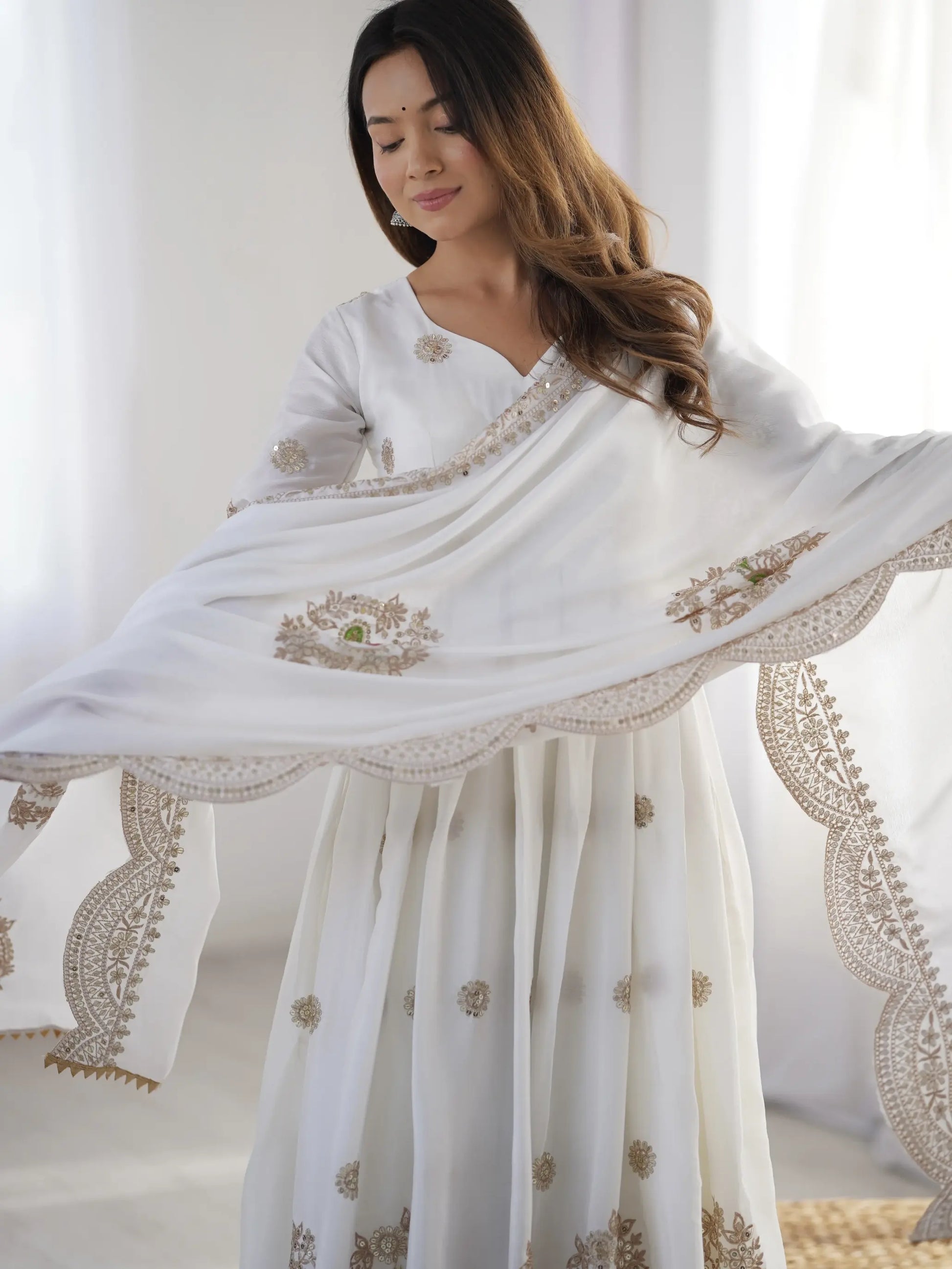 Woman holding a white saree with gold embroidery against a white curtain background