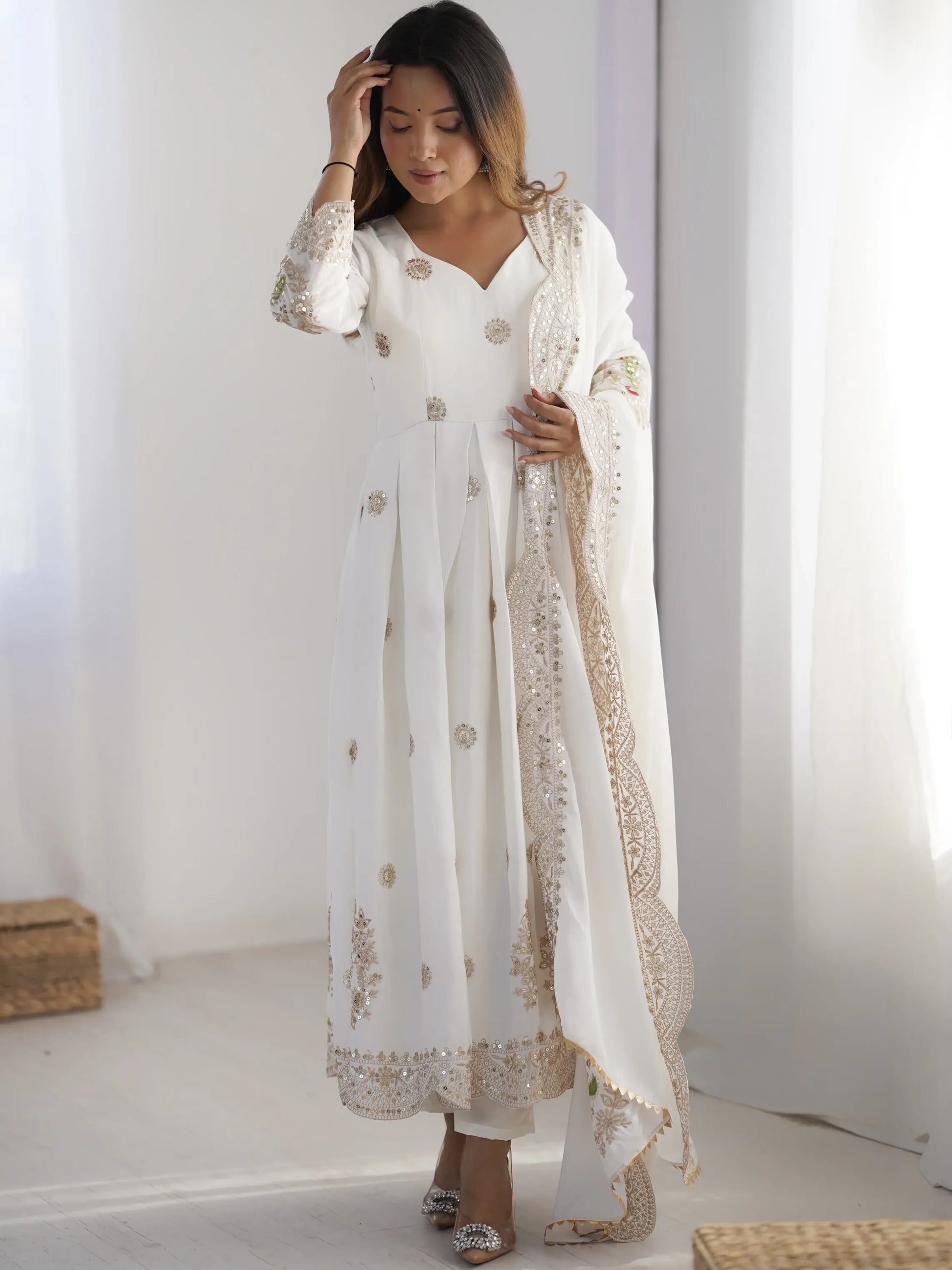 Woman wearing a white embroidered outfit with a sheer dupatta in a minimalistic setting.