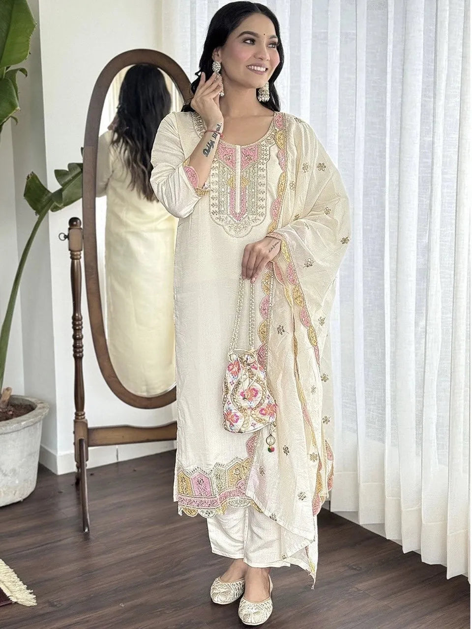 Woman in a white embroidered outfit standing in a room with a mirror and plant.