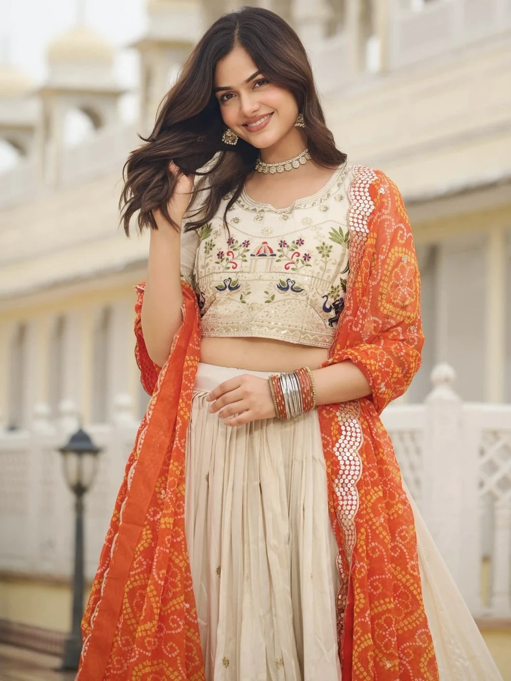Woman wearing an embroidered top and beige skirt with an orange dupatta, standing outdoors.