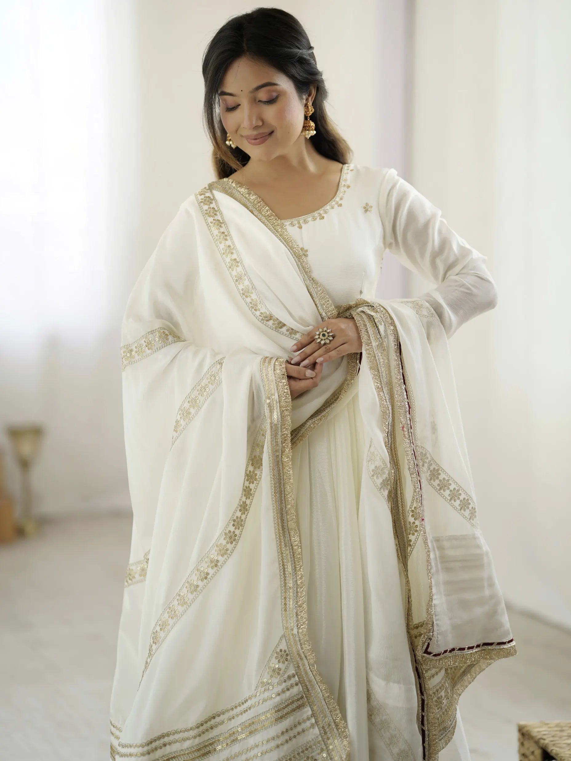 Woman wearing a white saree with gold embroidery in a bright room.