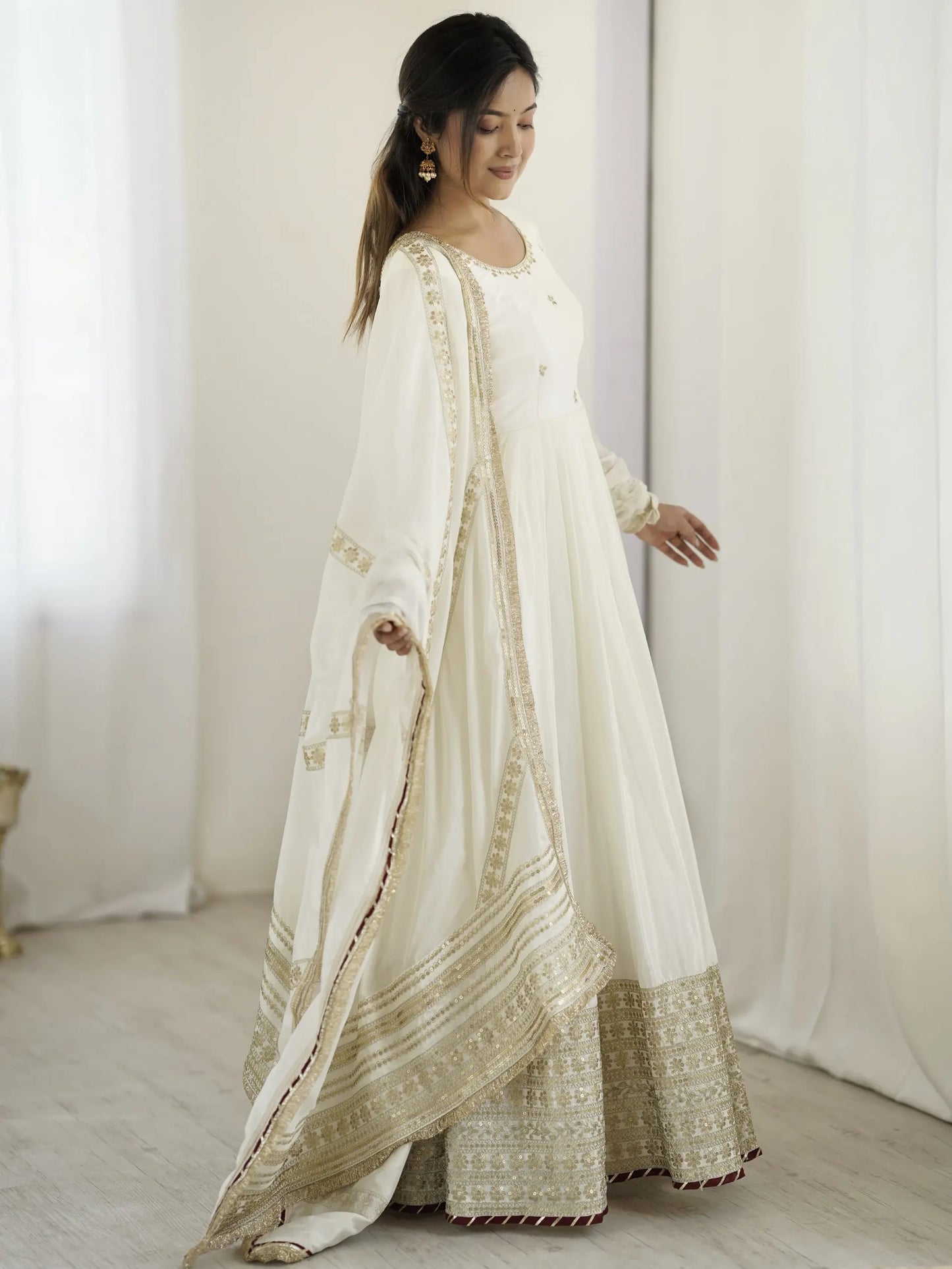 Woman wearing a traditional white and gold outfit with intricate patterns in a minimalistic setting.