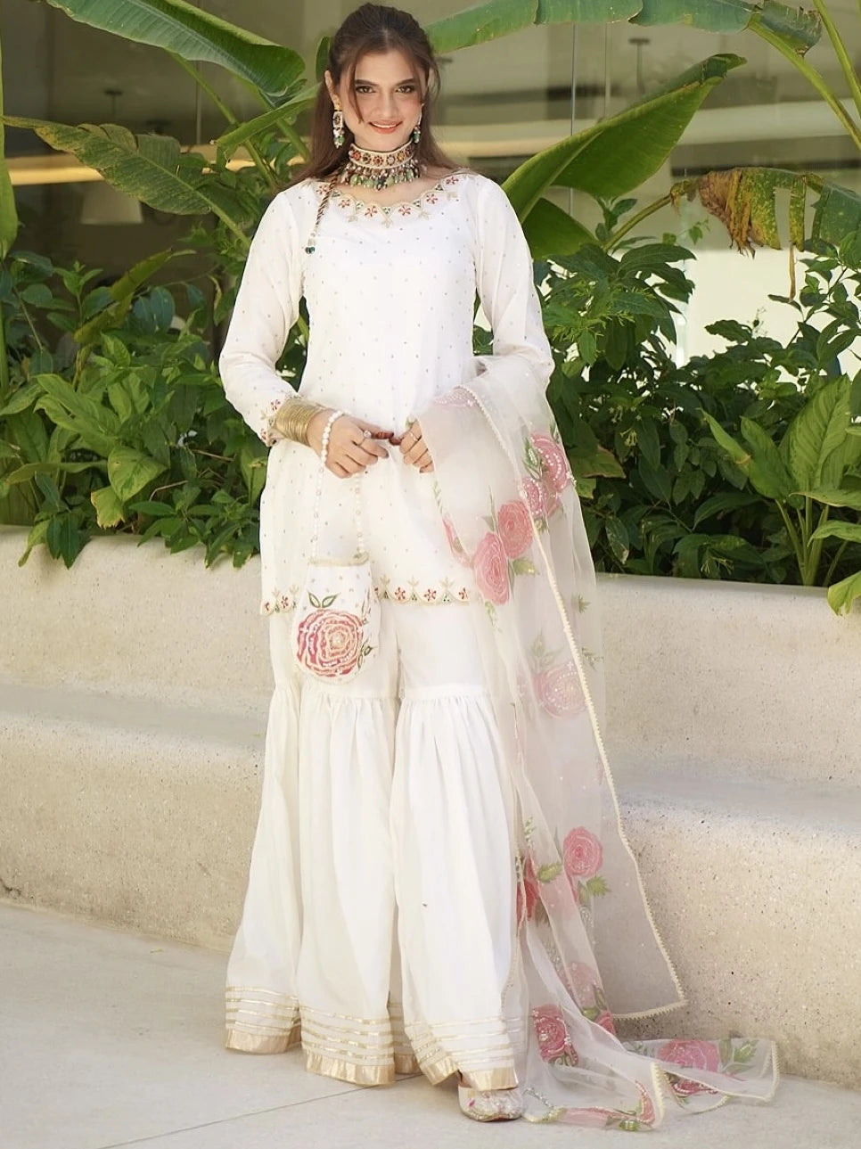 Woman in a white traditional outfit with floral details standing in front of green plants.