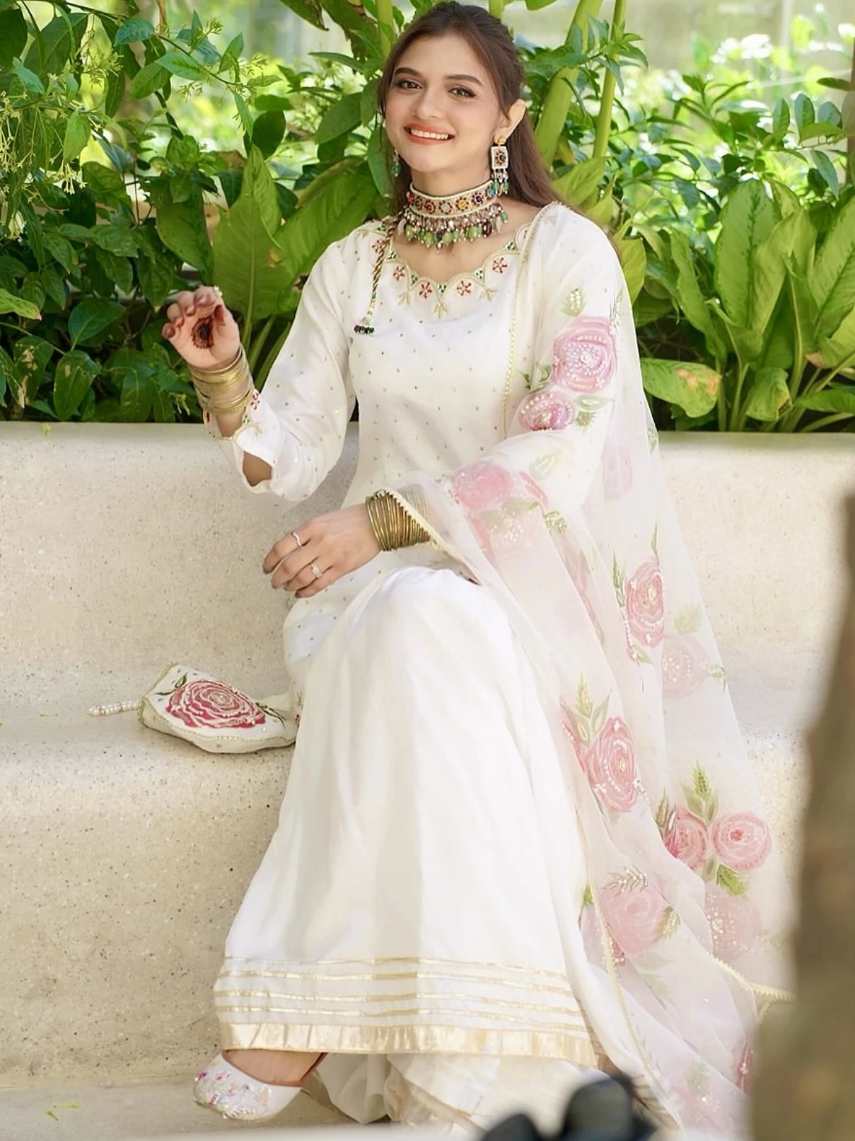 Woman in a white traditional outfit with floral patterns sitting on a couch with greenery in the background