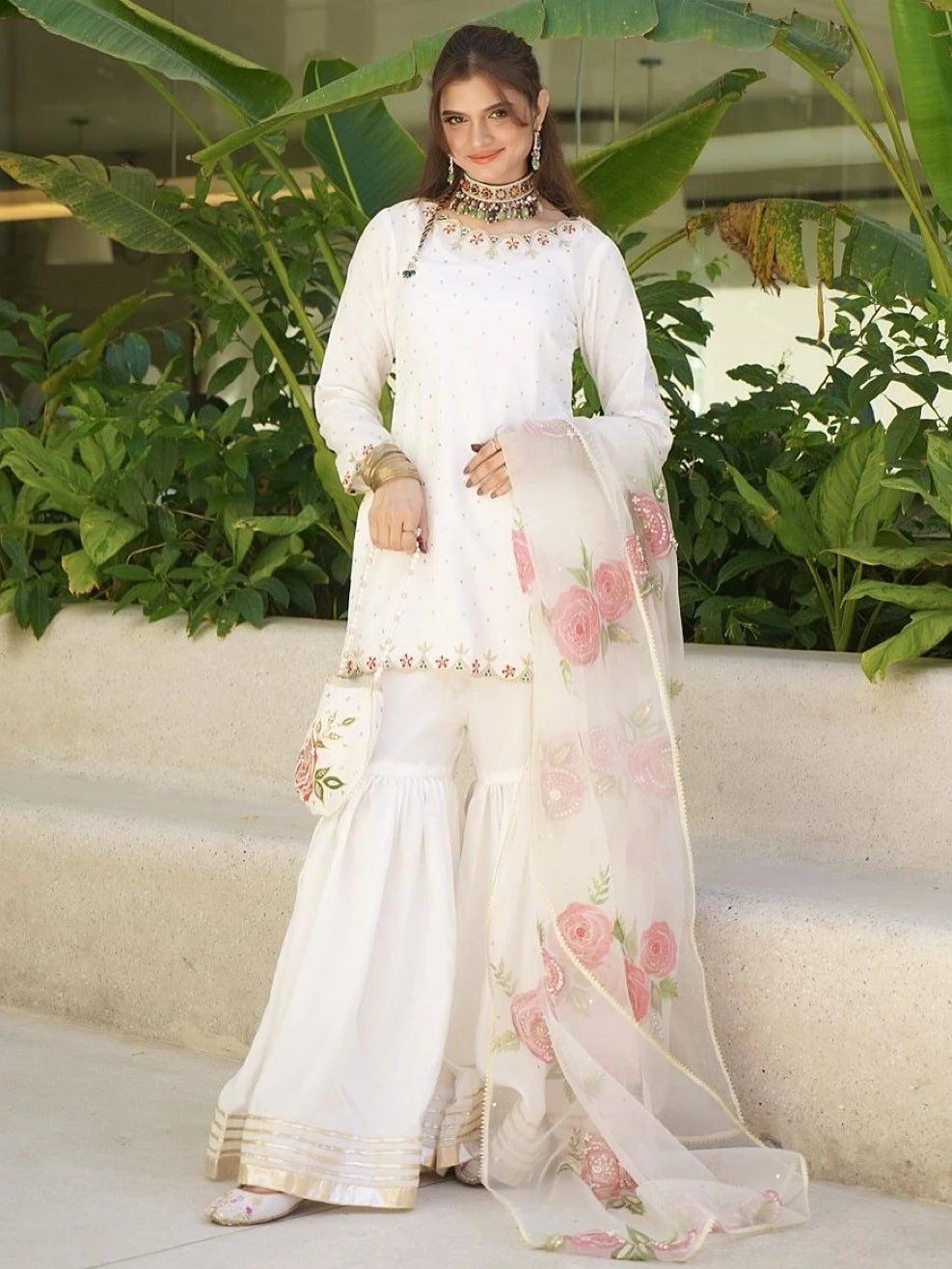 Woman in a white traditional outfit with floral details standing in front of green plants.