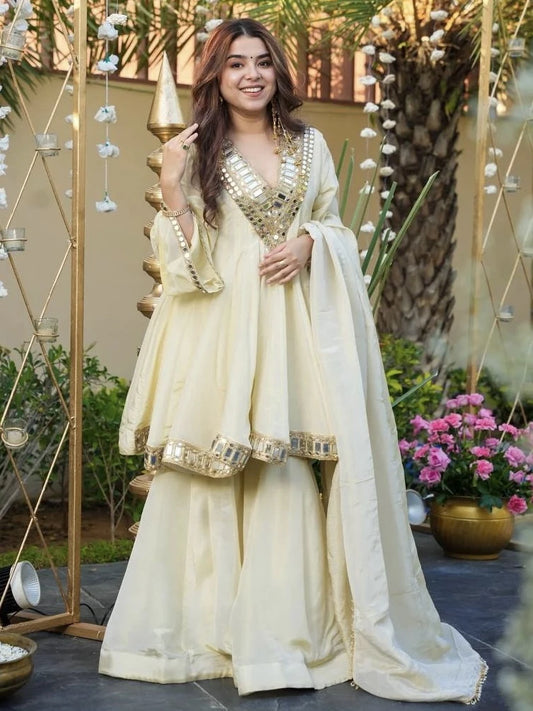 Woman in a traditional cream-colored outfit with gold embroidery standing outdoors.