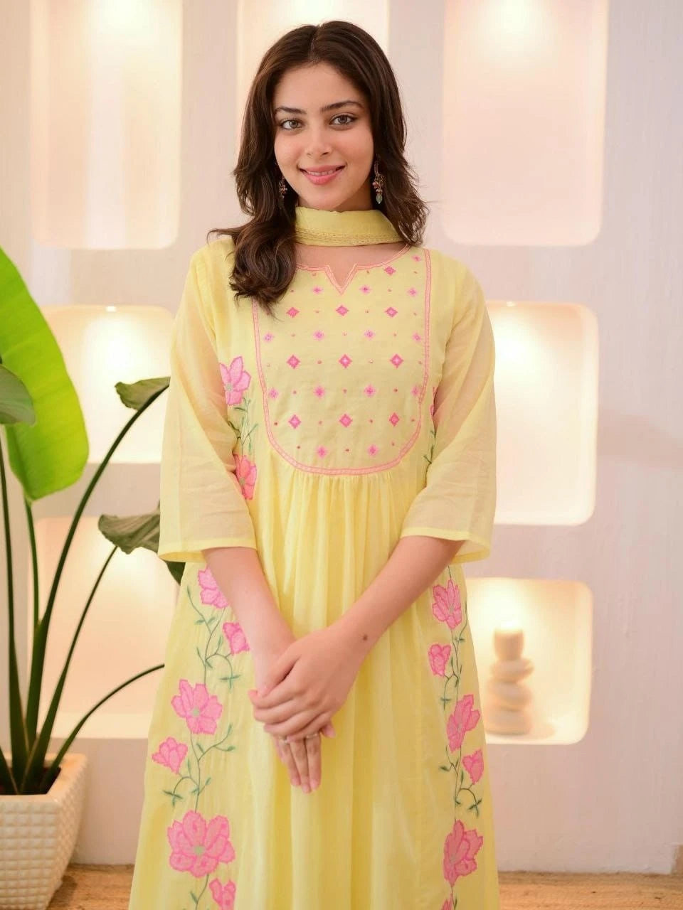 Woman wearing a light yellow dress with pink floral embroidery in a softly lit room.