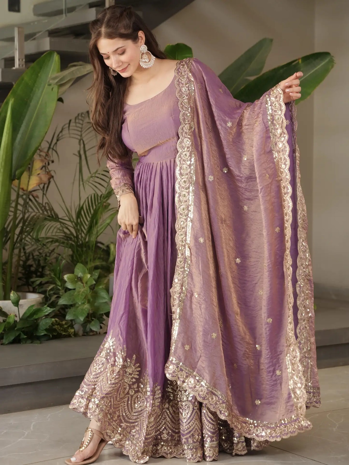 Woman in a purple traditional outfit with gold embroidery, standing indoors with plants in the background.
