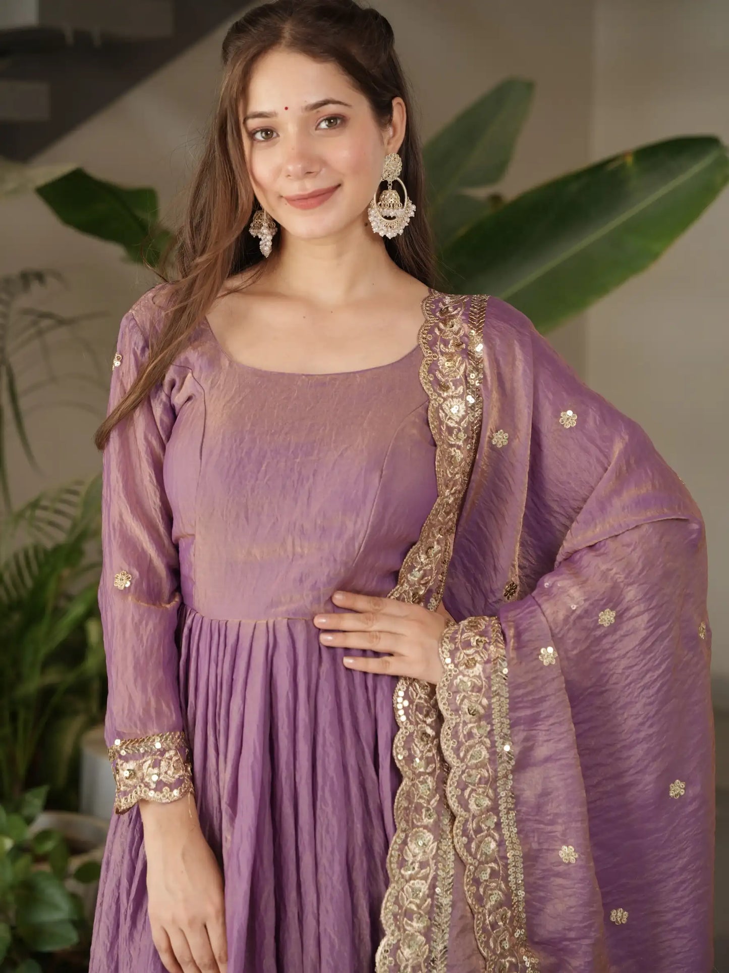 Woman wearing a purple traditional outfit with gold embroidery, standing in front of green plants.