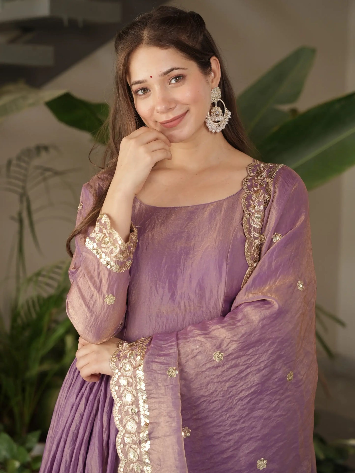 Woman wearing a purple traditional outfit with gold embroidery, standing against a blurred indoor background.