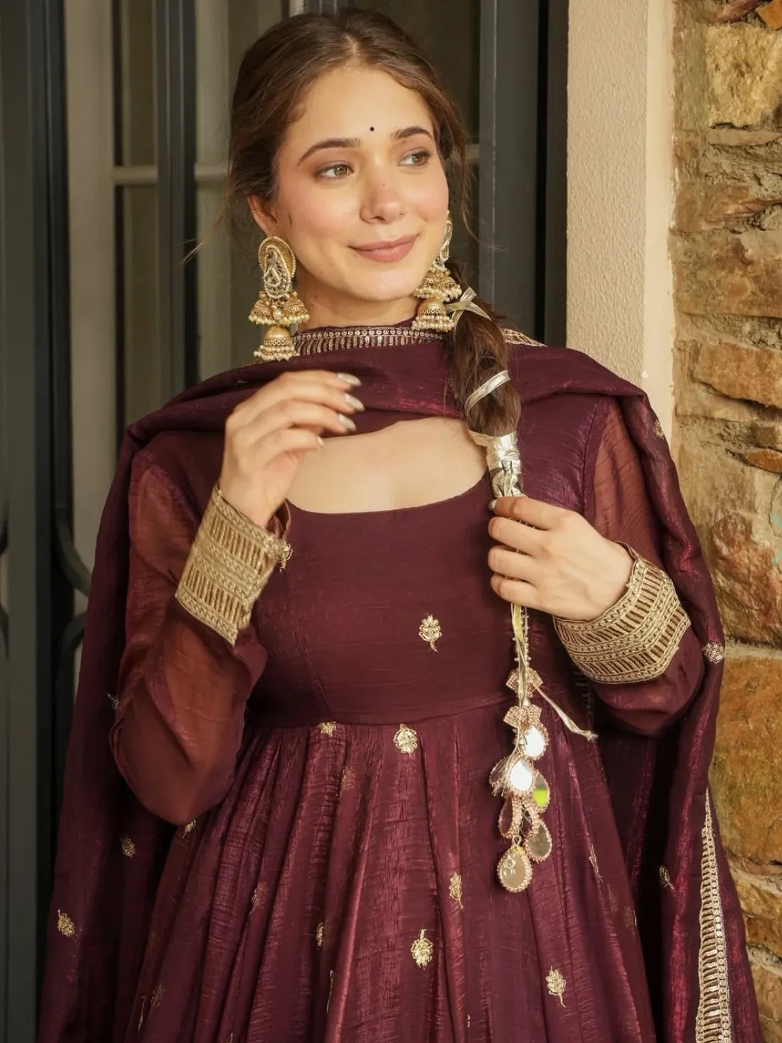 Woman in traditional maroon outfit with gold accents holding a decorative item, standing against a stone wall.