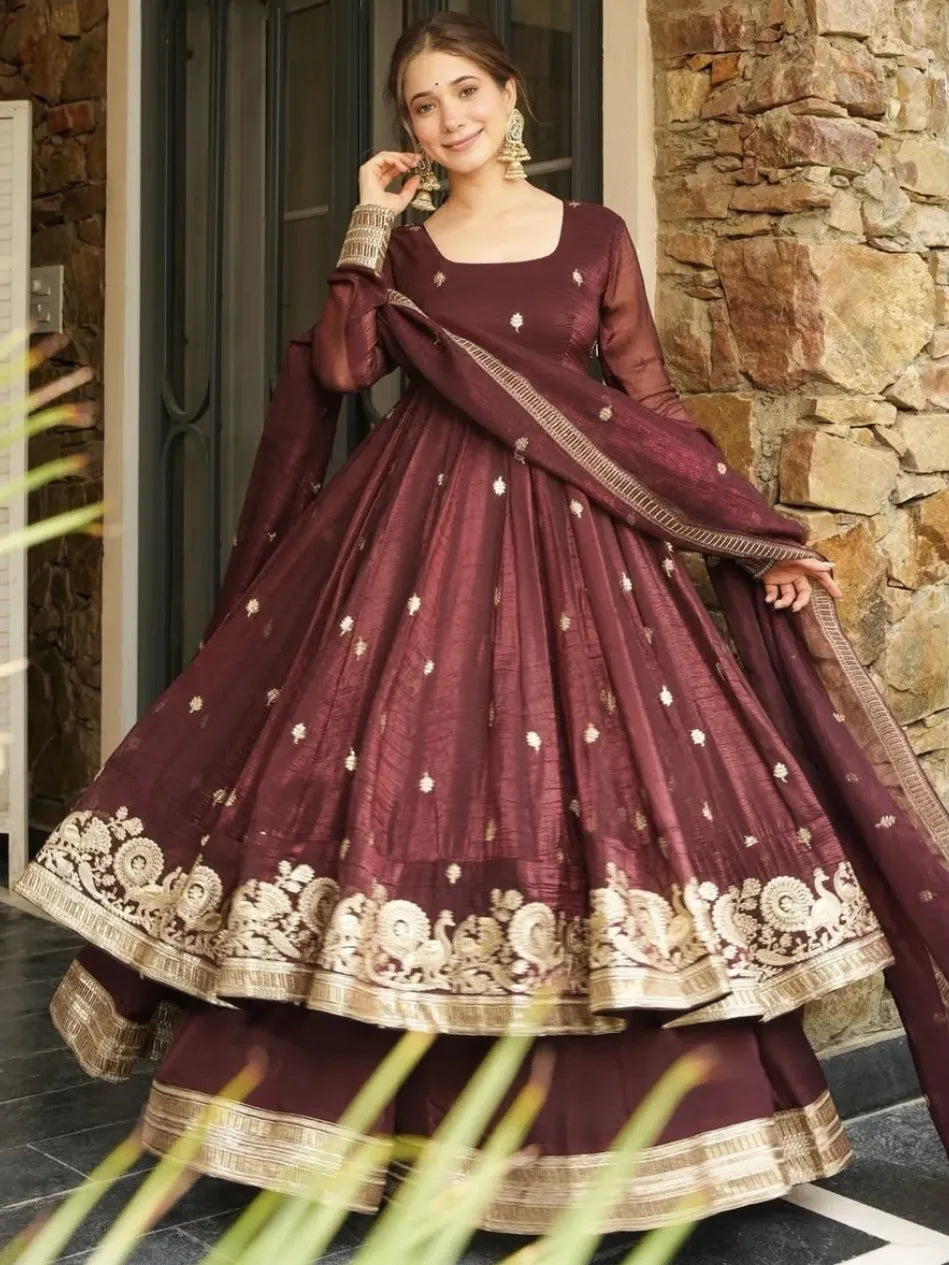 Woman wearing a maroon traditional outfit with gold embroidery against a stone wall.