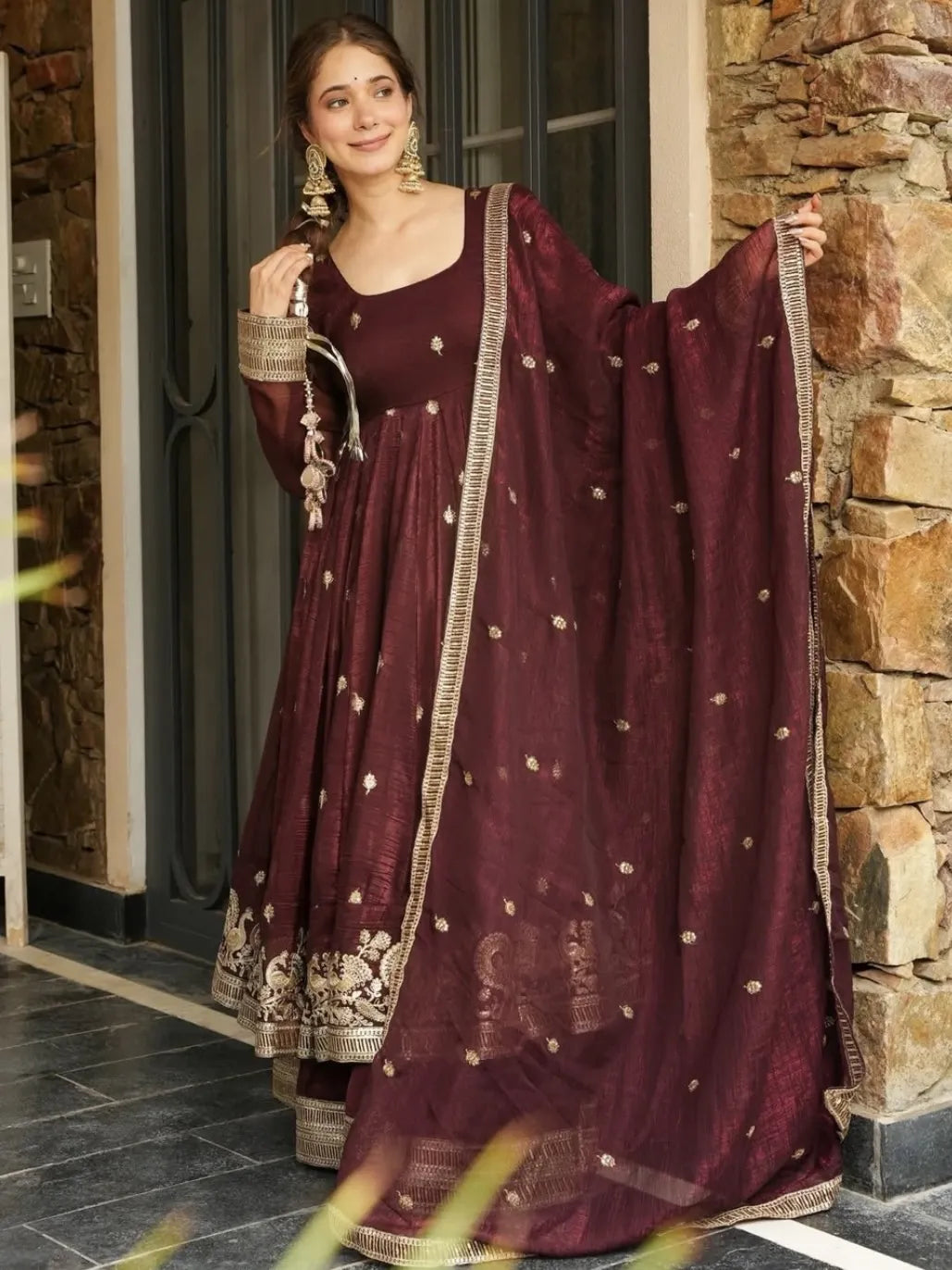 Woman in a maroon traditional outfit with gold details standing in front of a stone wall.