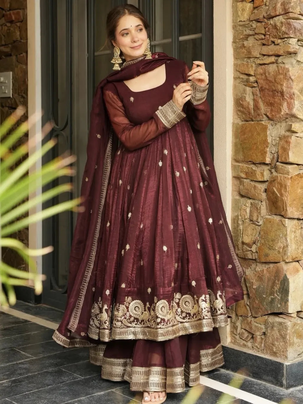 Woman in a maroon traditional outfit with gold embroidery standing in front of a stone wall.