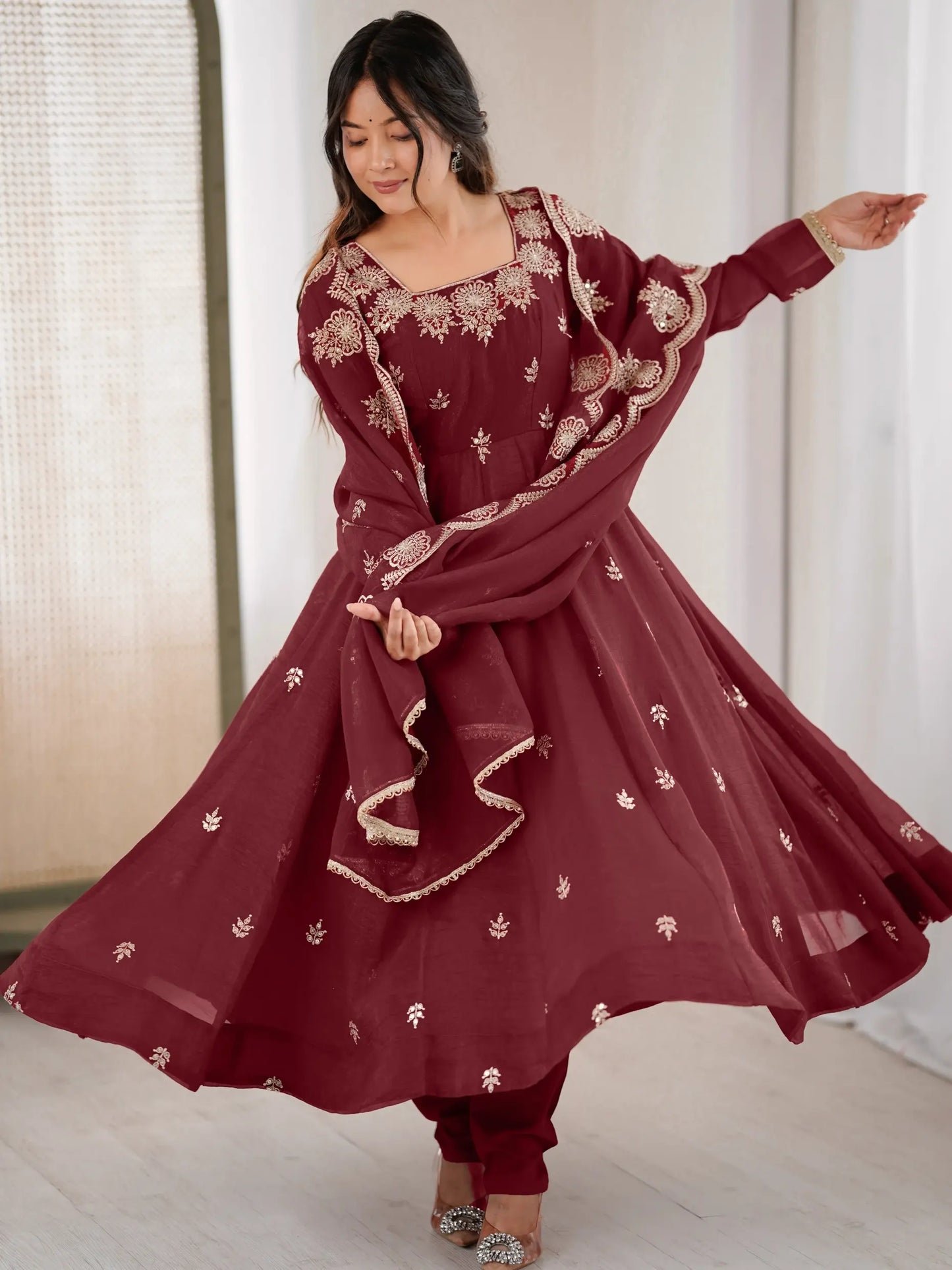 Woman in a maroon traditional outfit with white patterns, standing indoors.