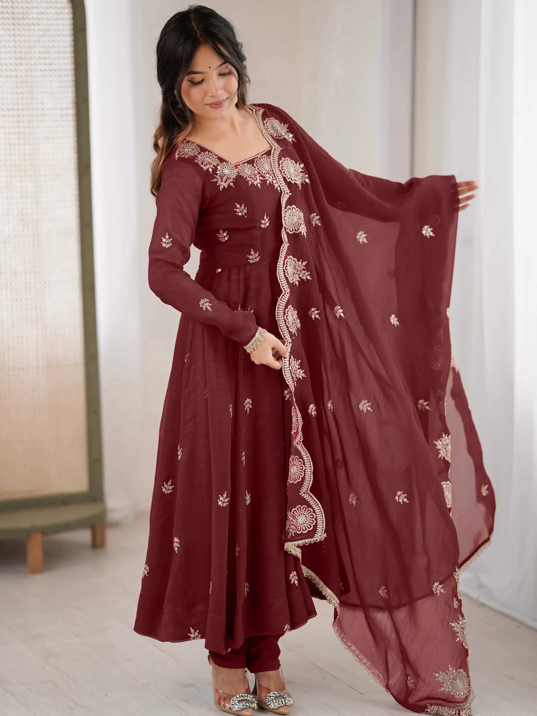 Woman wearing a maroon traditional outfit with white embroidery in a minimal indoor setting.