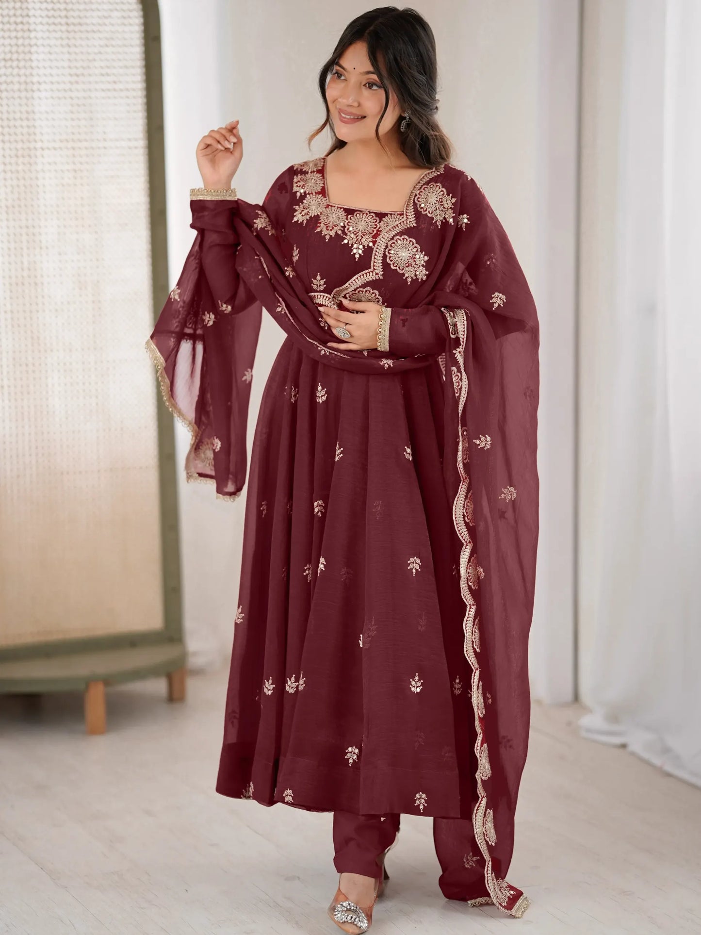 Woman wearing a maroon traditional outfit with white embroidery in a minimal indoor setting.