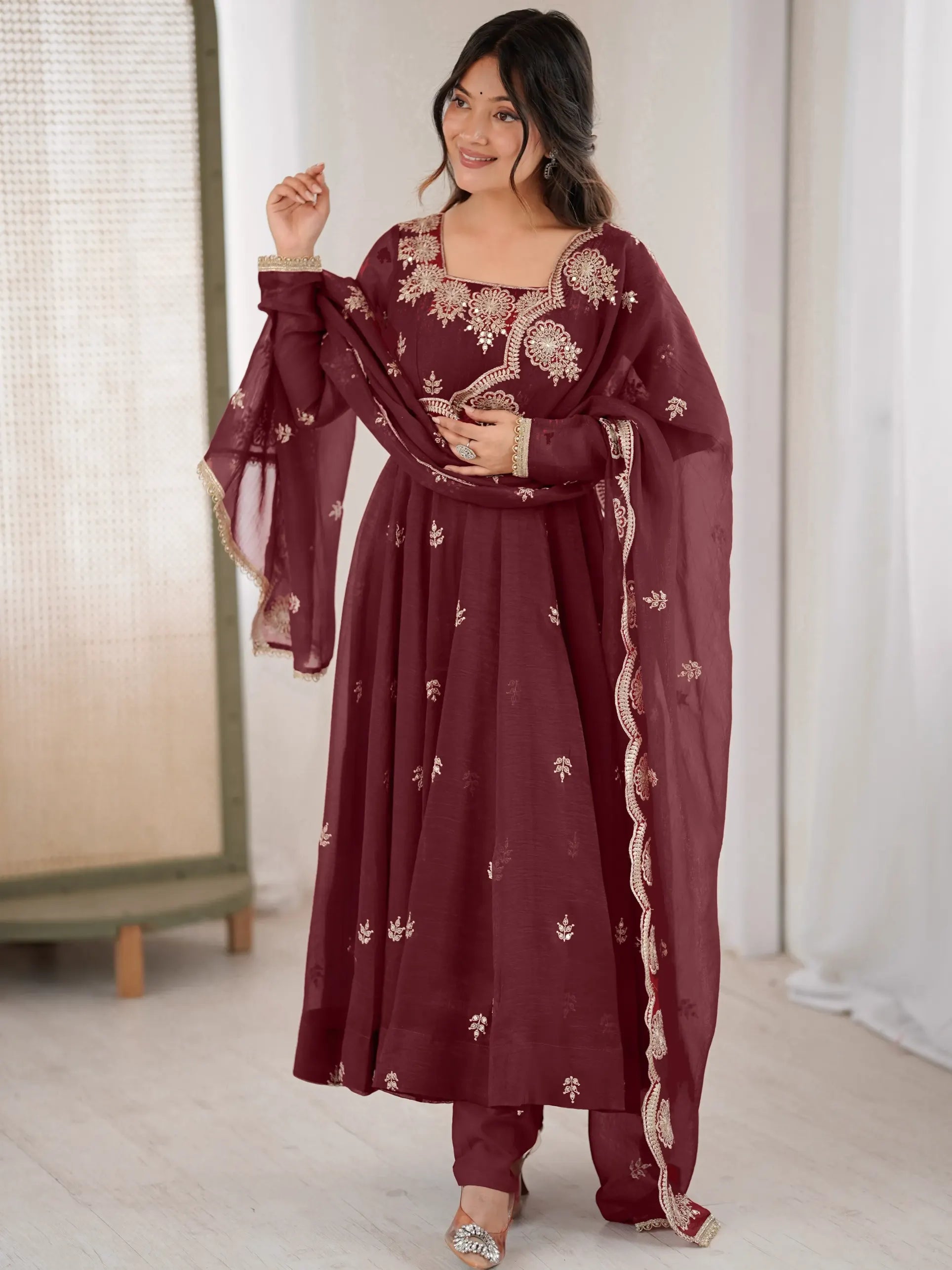 Woman wearing a maroon traditional outfit with white embroidery in a minimal indoor setting.