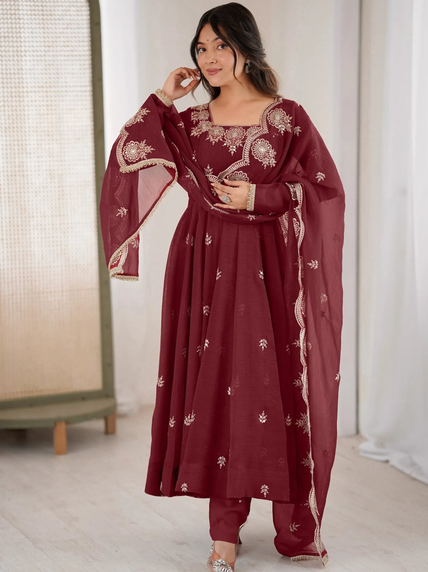Woman wearing a maroon traditional outfit with gold embroidery in a minimal indoor setting.