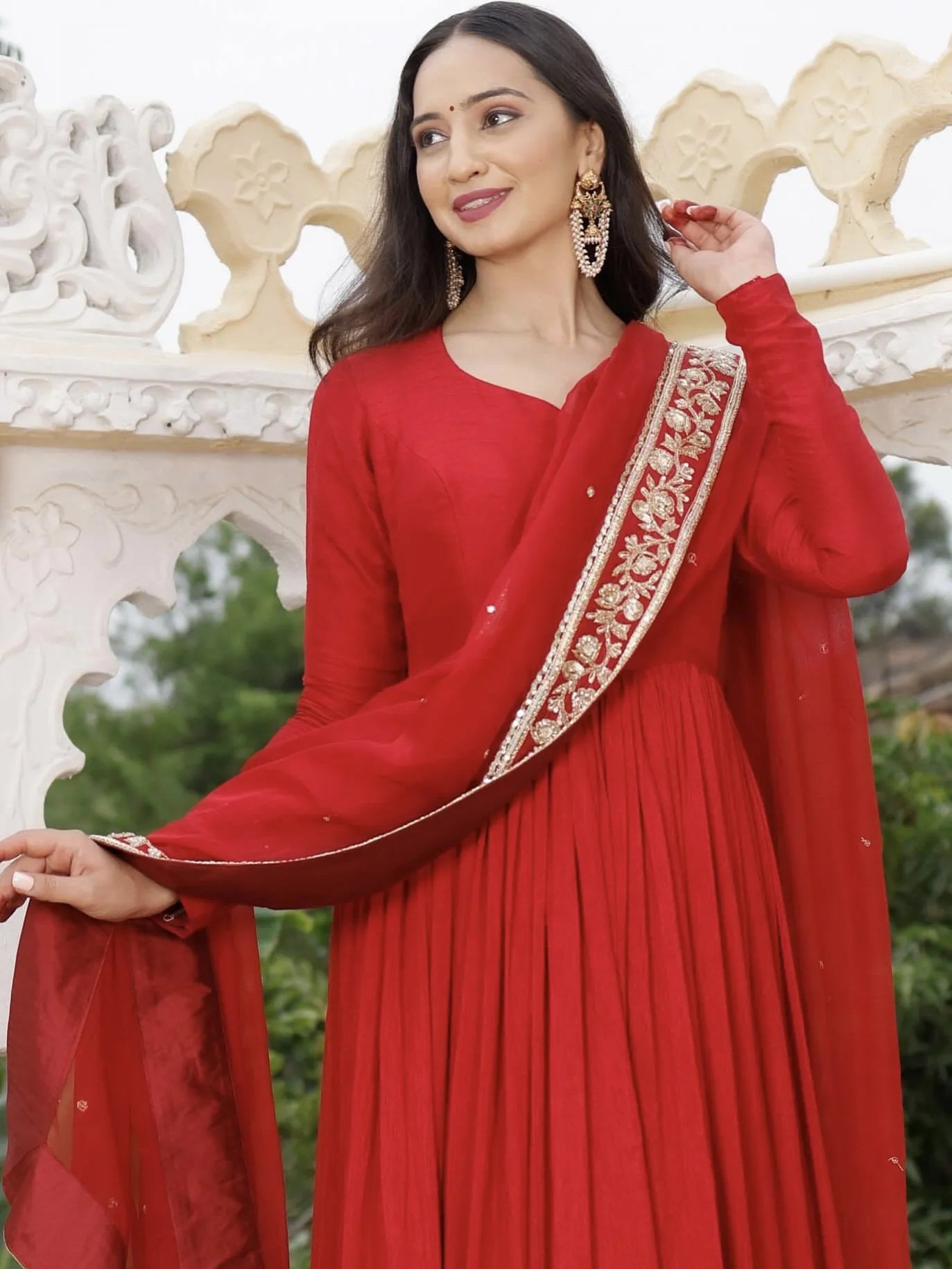 Woman in a red traditional outfit with a decorative border, standing against a white architectural background.