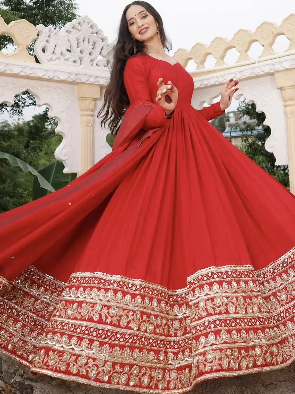 Woman in a red traditional outfit with gold embroidery standing in front of decorative stone architecture.