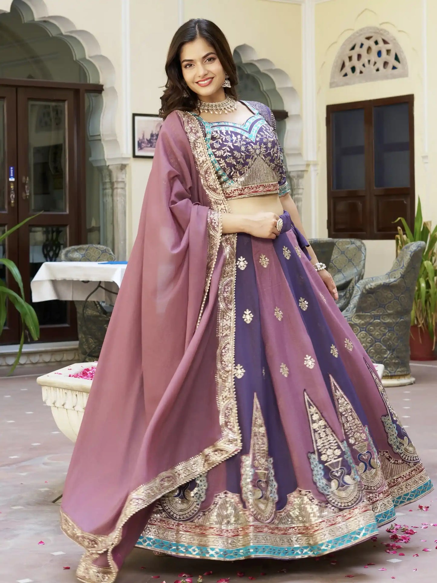 Woman in a traditional outfit with a purple and pink color scheme standing in an elegant indoor setting.