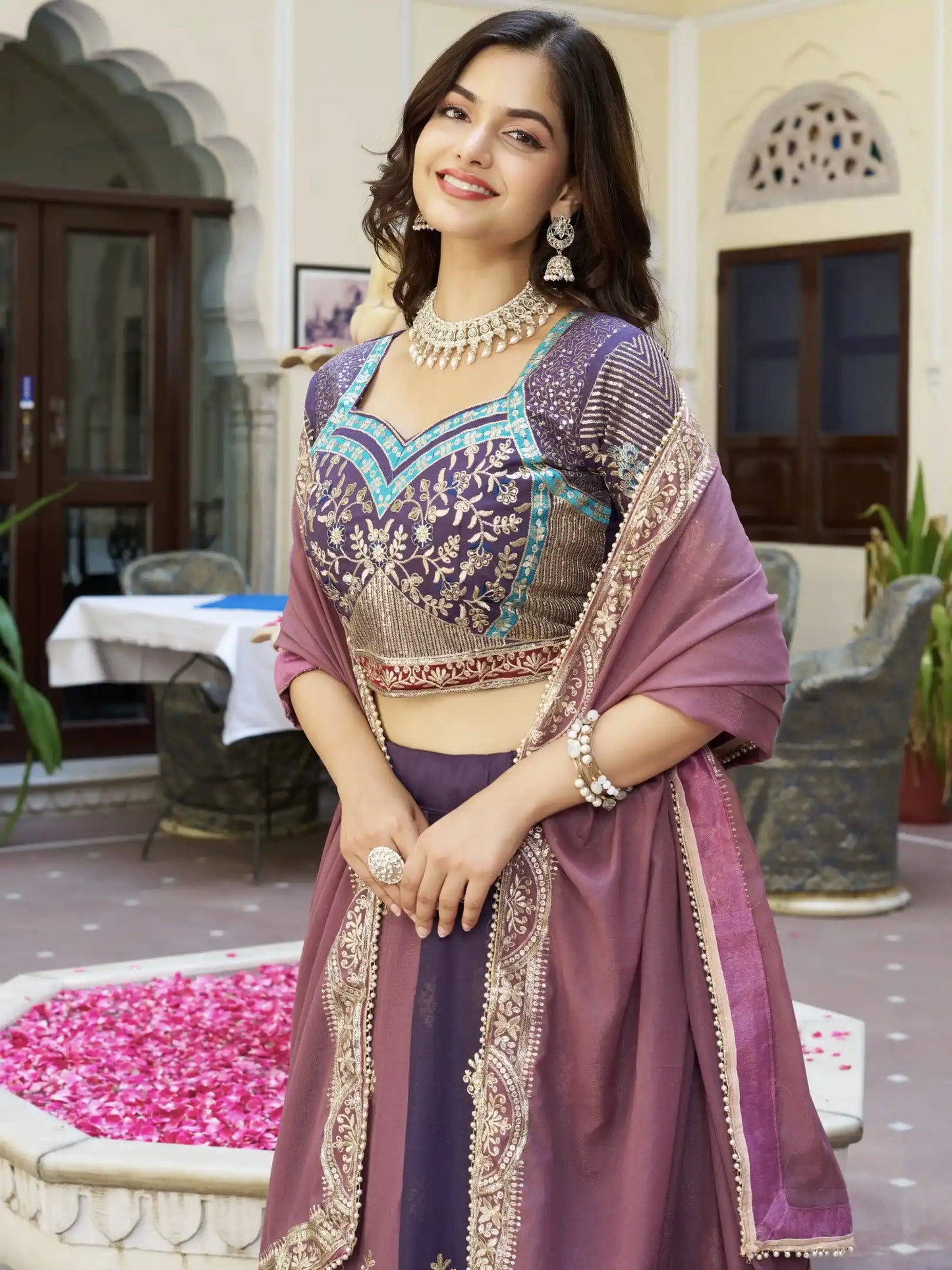 Woman in a traditional purple and gold outfit standing in an outdoor setting with decorative elements.
