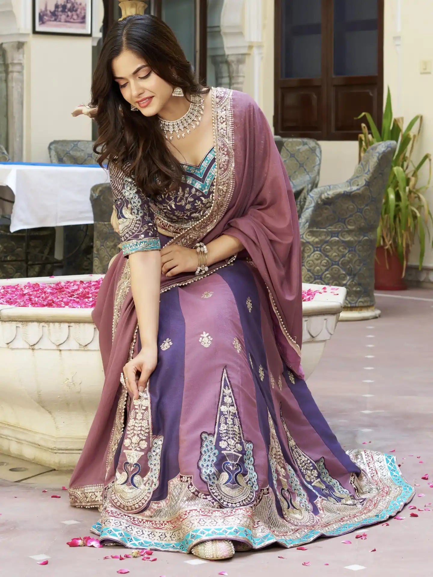 Woman in a traditional purple and blue embroidered saree with jewelry, standing outdoors.
