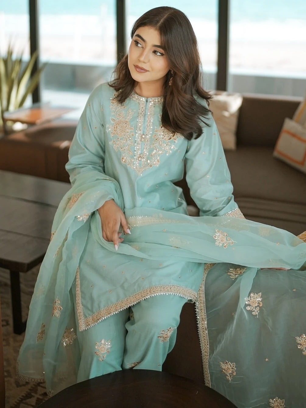 Woman in a light blue embroidered traditional outfit sitting indoors.