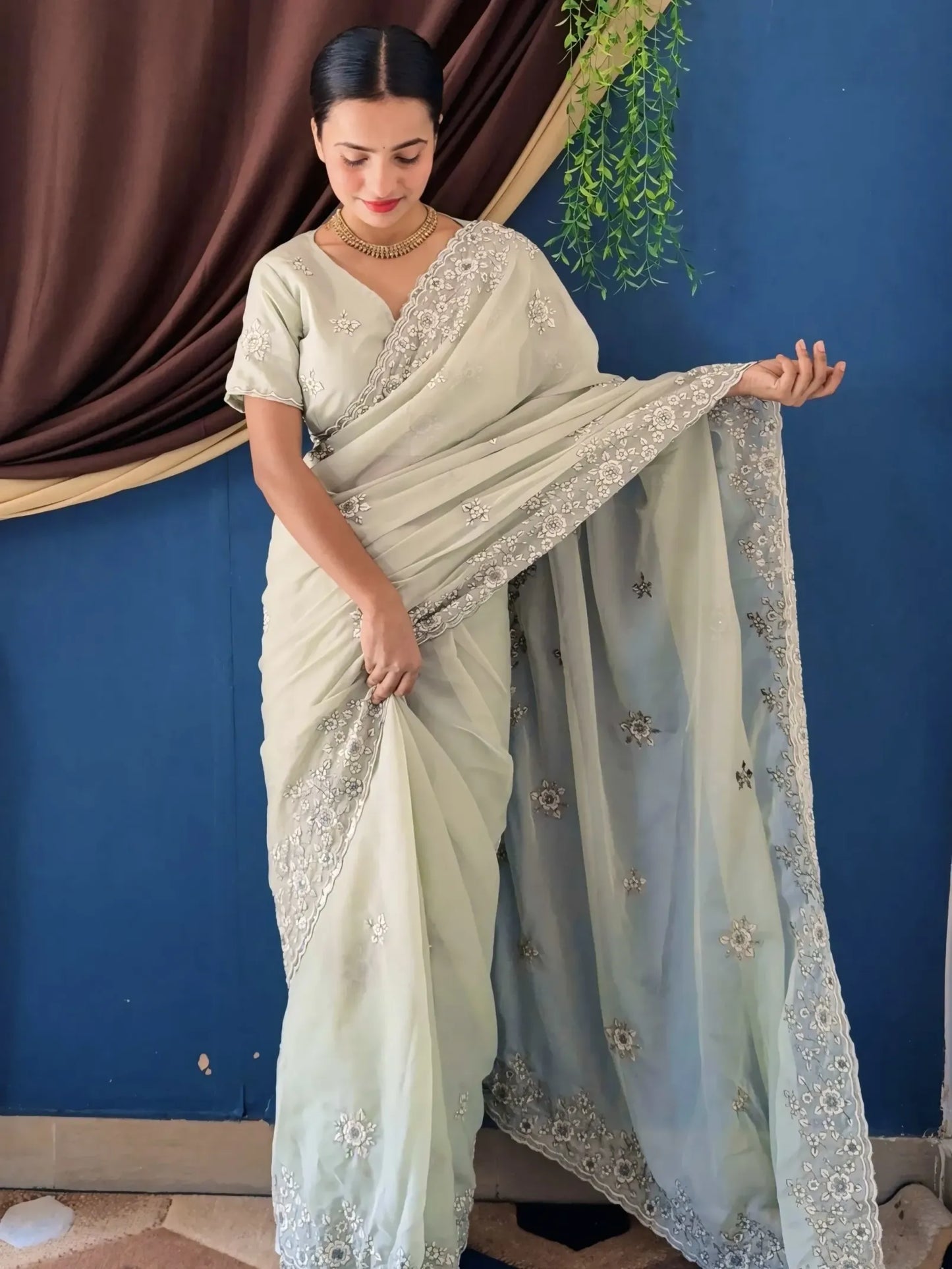 Woman wearing a light green saree with silver embroidery against a blue wall.