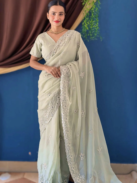 Woman wearing a light green saree with white embroidery against a blue wall.