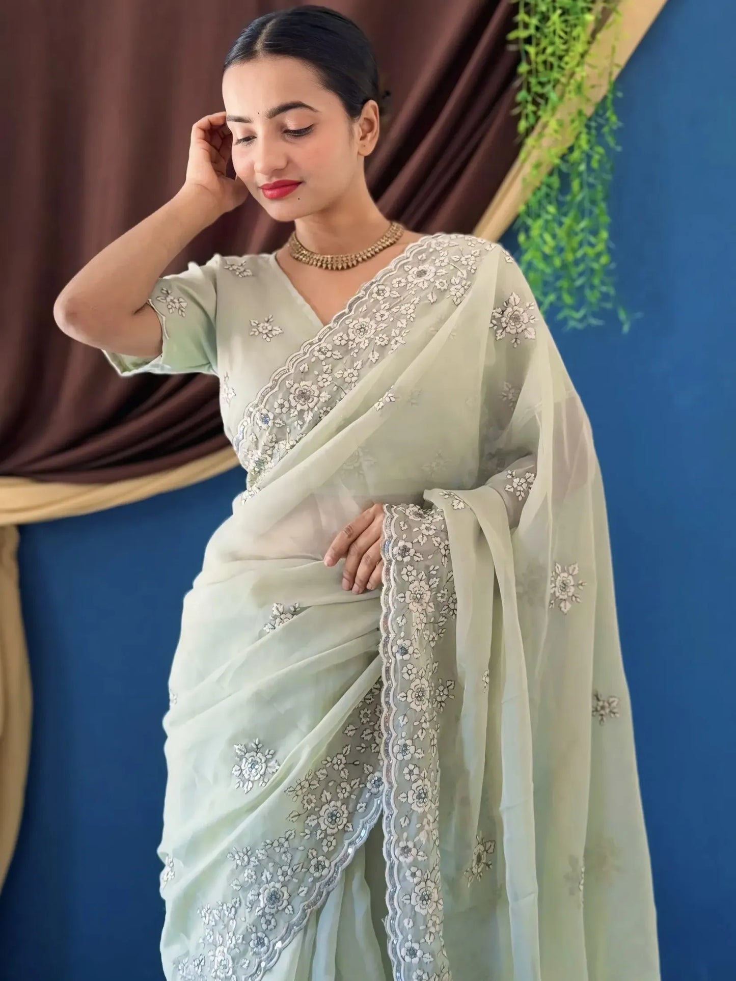 Woman wearing a light green saree with white embroidery against a blue and brown curtain background