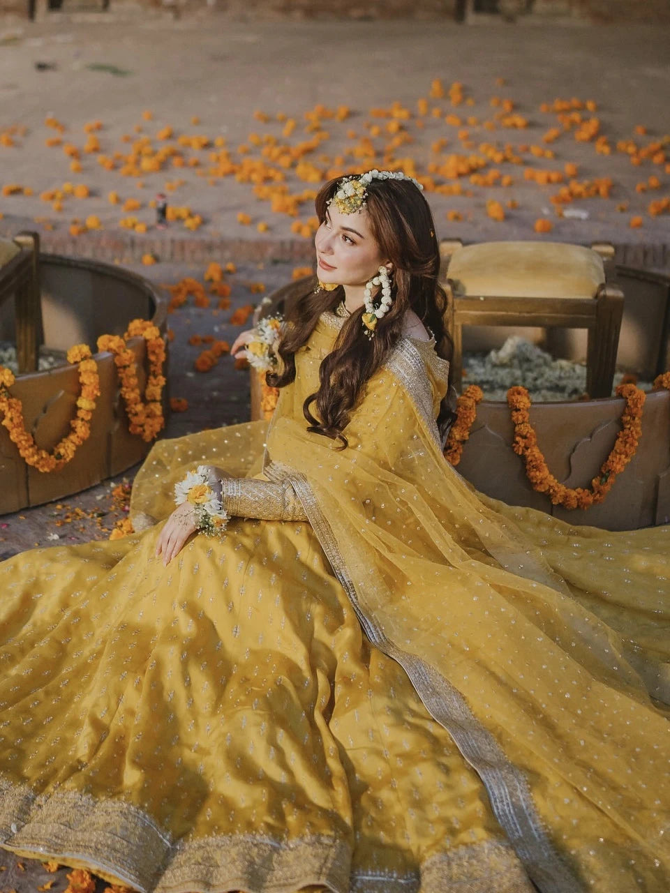 Woman in a yellow traditional outfit with floral decorations in an outdoor setting with orange flowers on the ground.