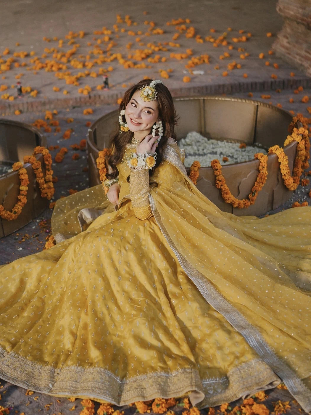 Woman in a yellow traditional outfit surrounded by orange flowers on a stone surface.