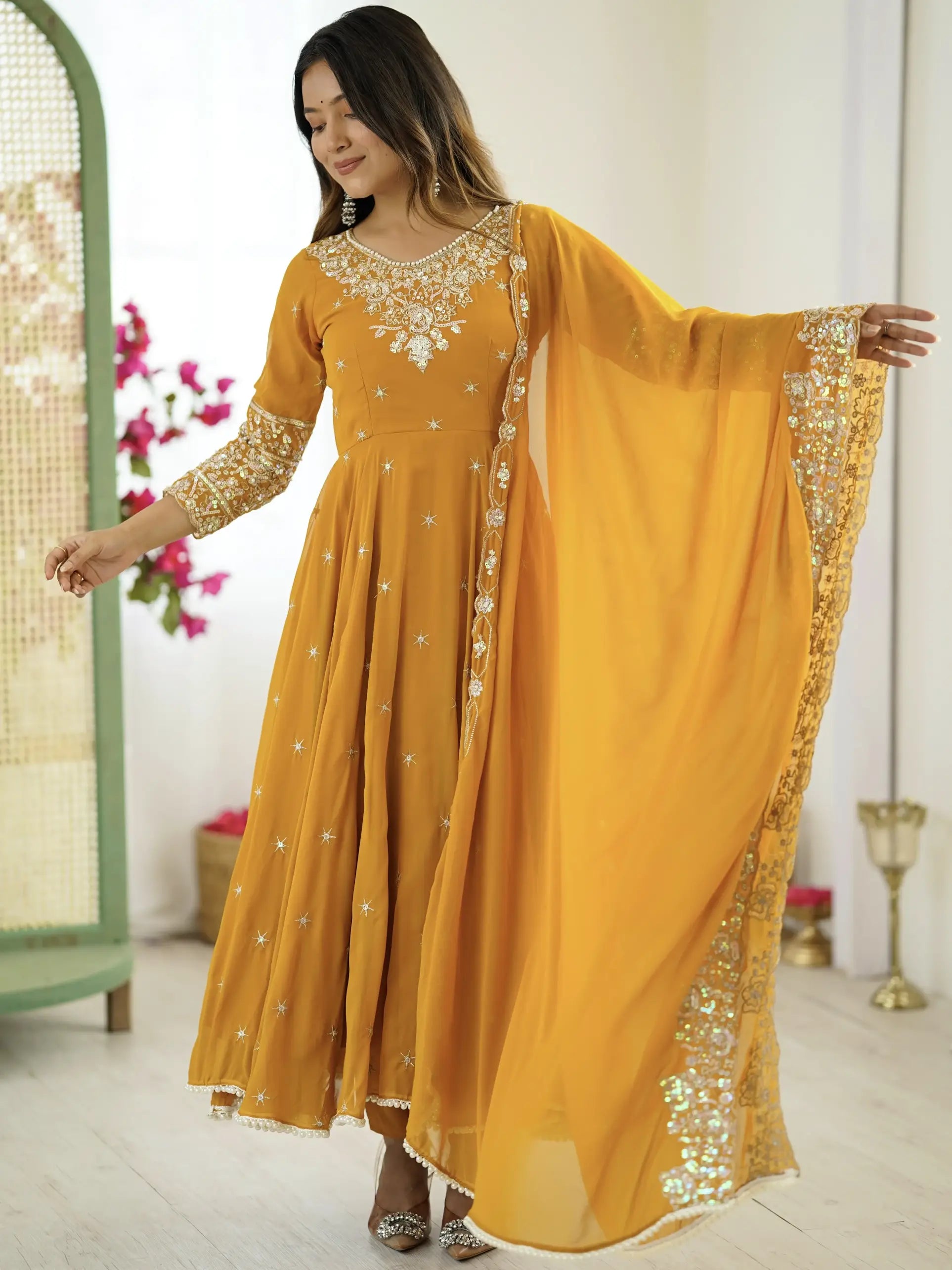 Woman wearing a yellow embroidered dress with a dupatta in a decorated room.