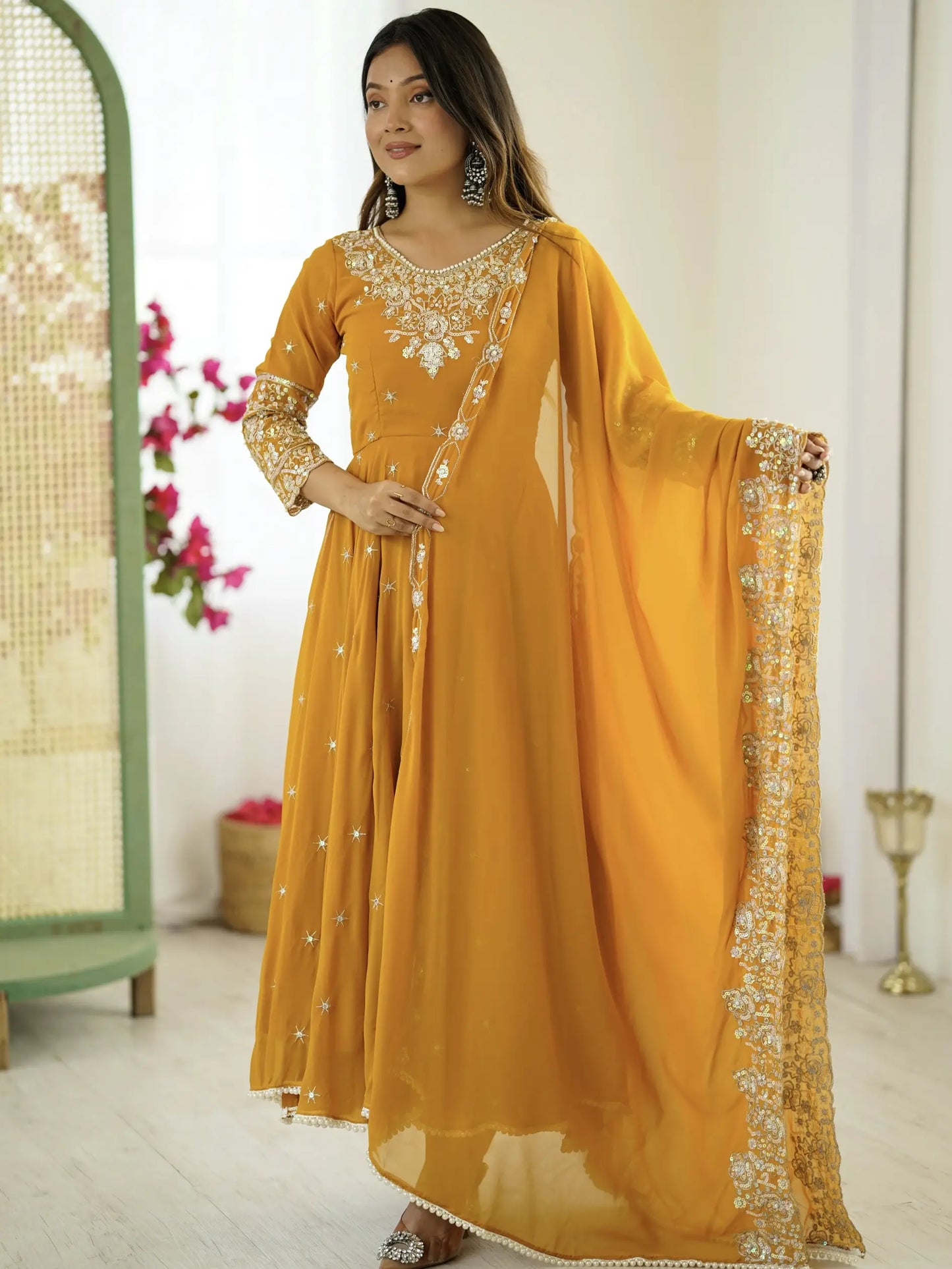 Woman wearing a mustard yellow dress with intricate white embroidery in a bright room.