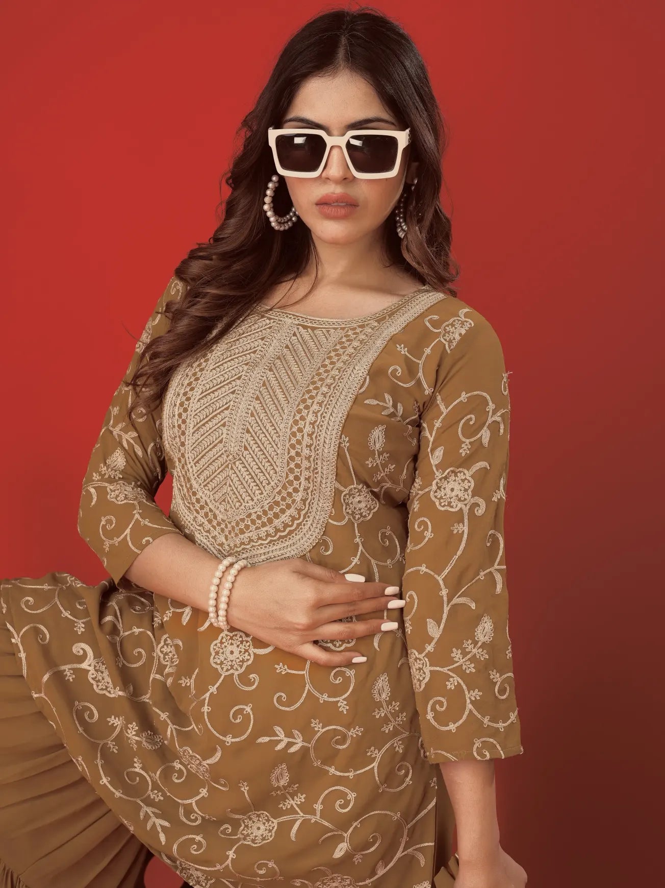 Woman wearing a brown embroidered traditional outfit with sunglasses against a red background