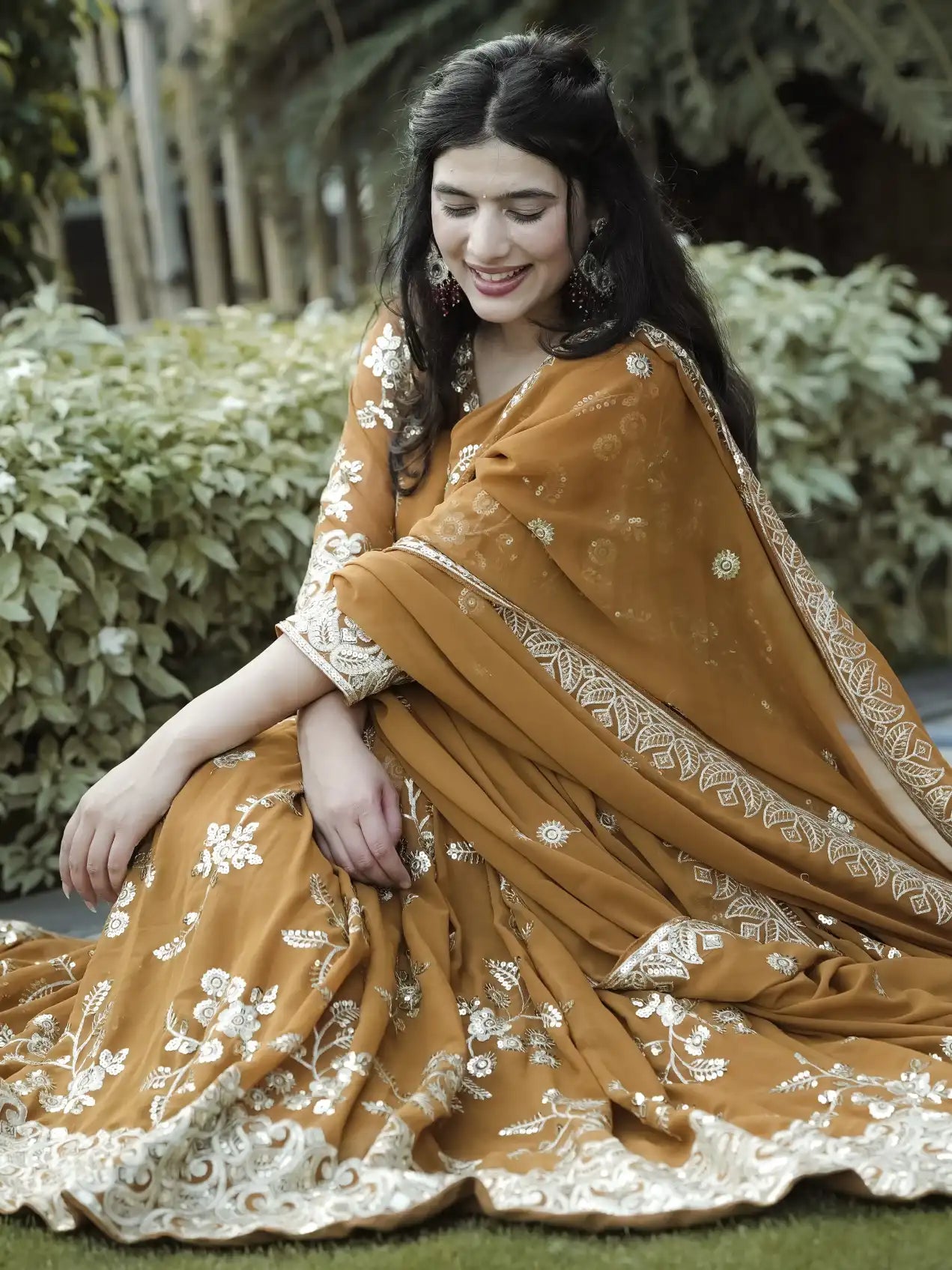 Woman in a traditional brown and gold saree sitting outdoors.