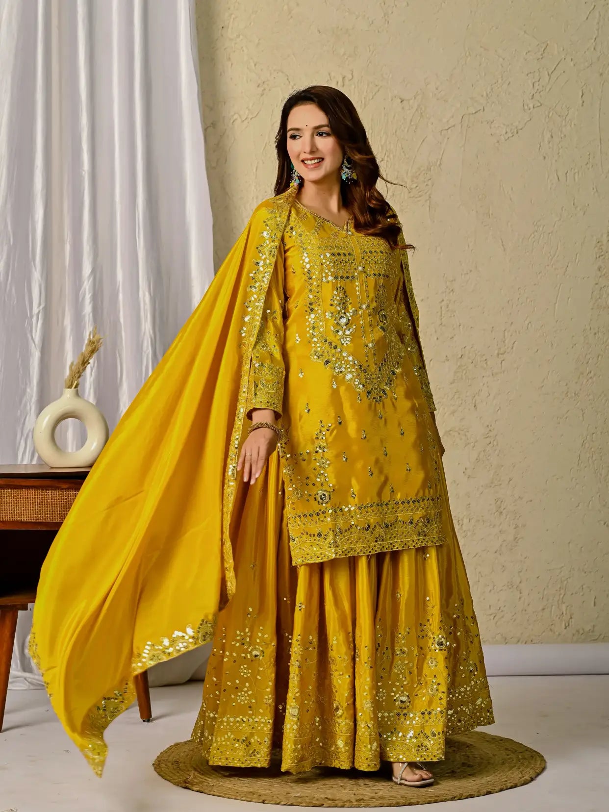 Woman wearing a yellow embroidered traditional outfit with a dupatta in a room setting.