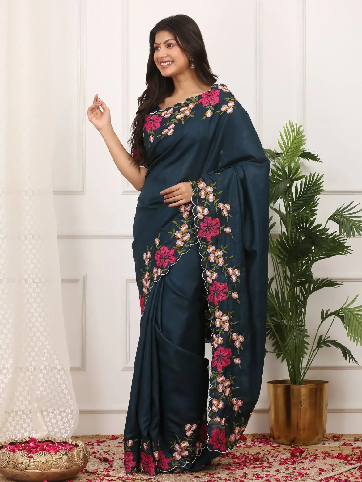 Woman wearing a dark blue saree with floral patterns in a room with a plant and decorative items.