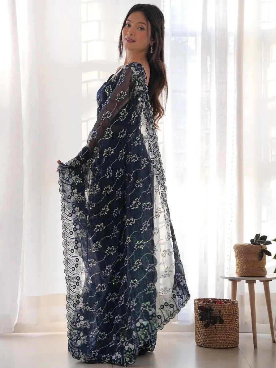 Woman wearing a dark blue floral dress standing in a bright room with white curtains.