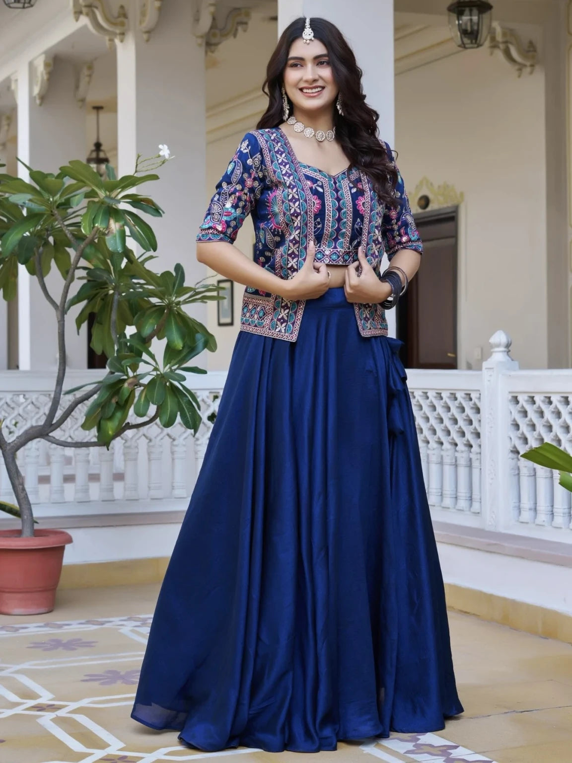 Woman in a traditional outfit with a floral top and blue skirt standing in an elegant indoor setting.