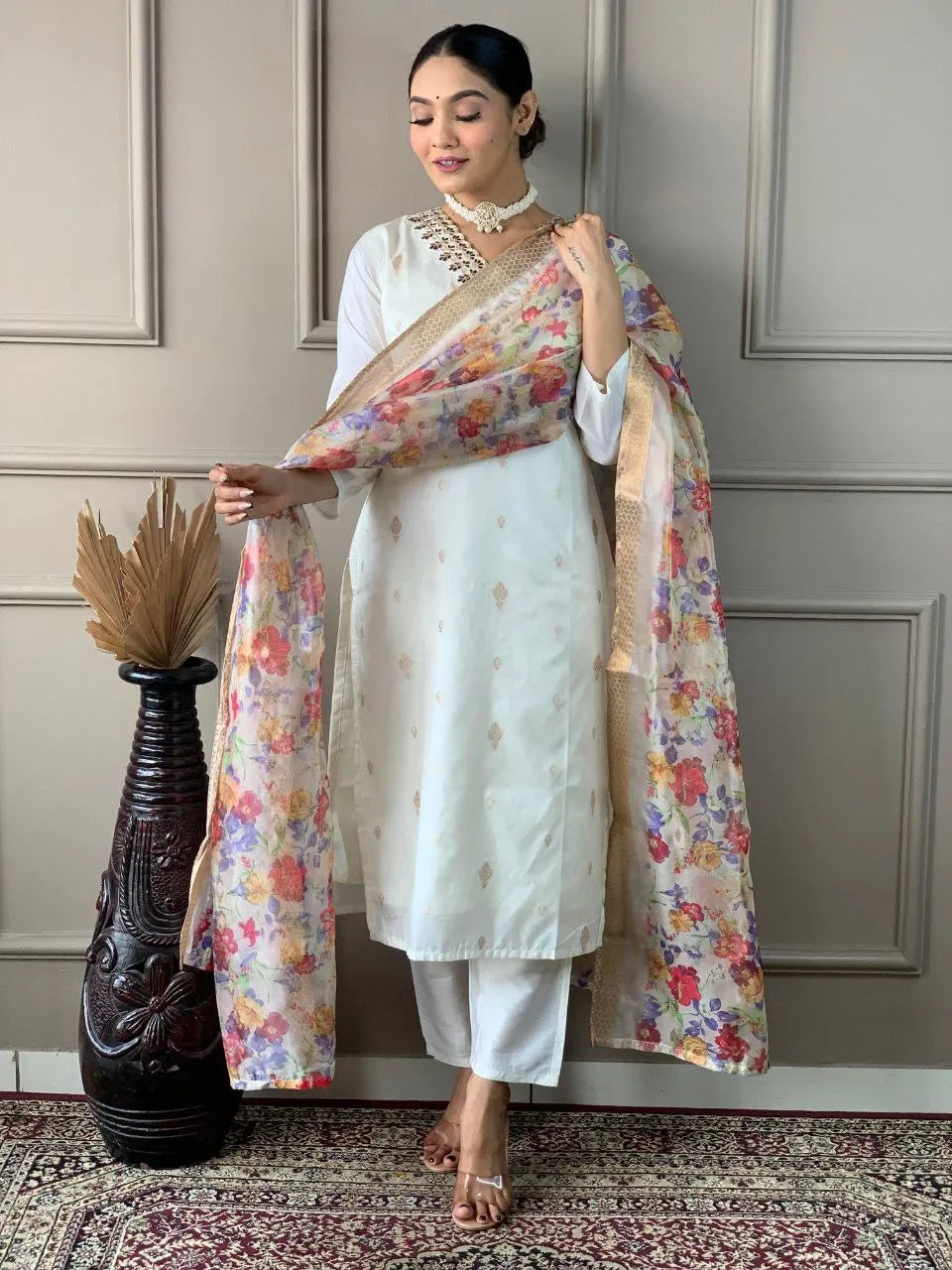 Woman in a white traditional outfit with a floral dupatta against a neutral wall.