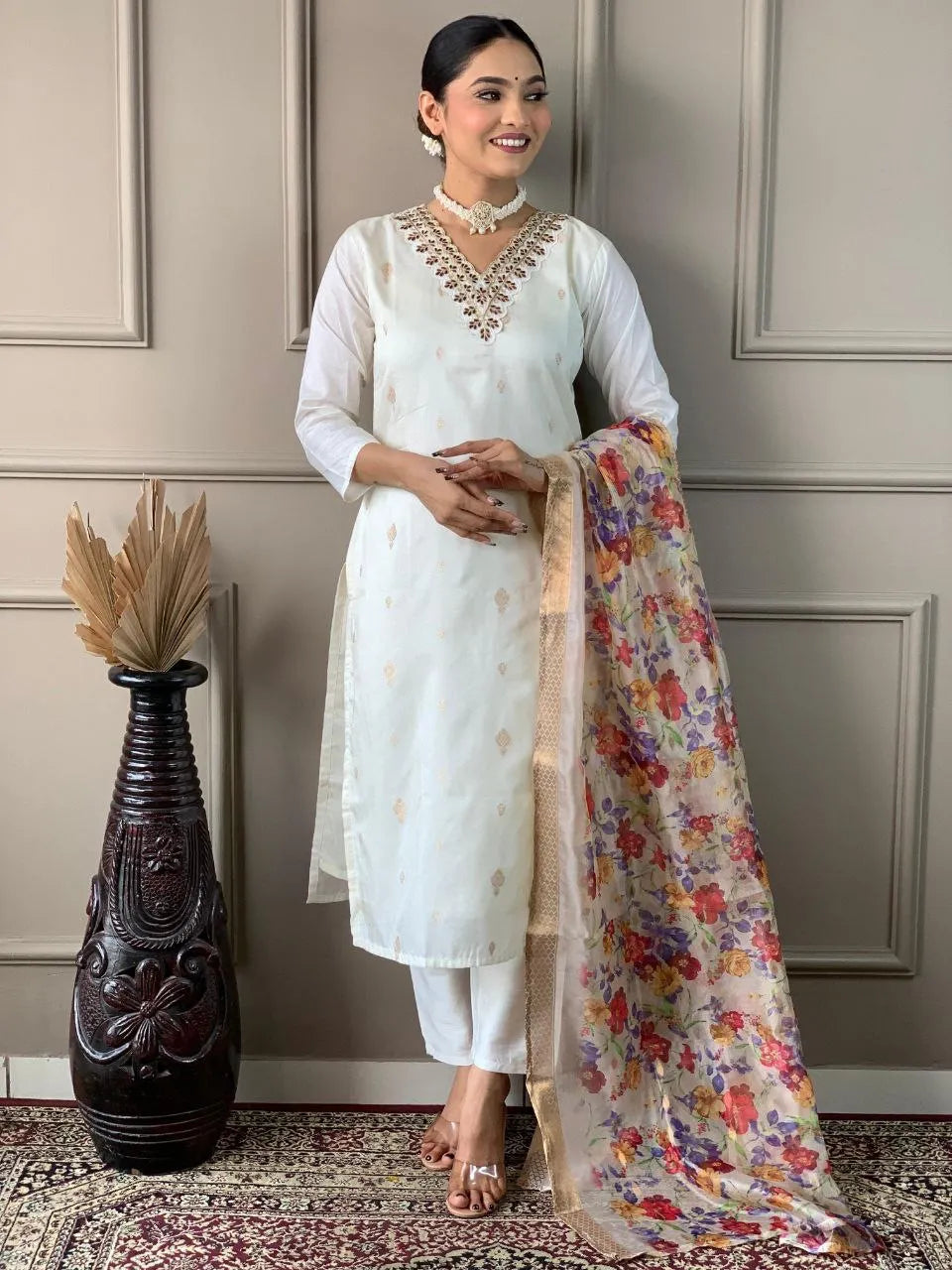 Woman in a white traditional outfit with a floral dupatta standing against a neutral wall.