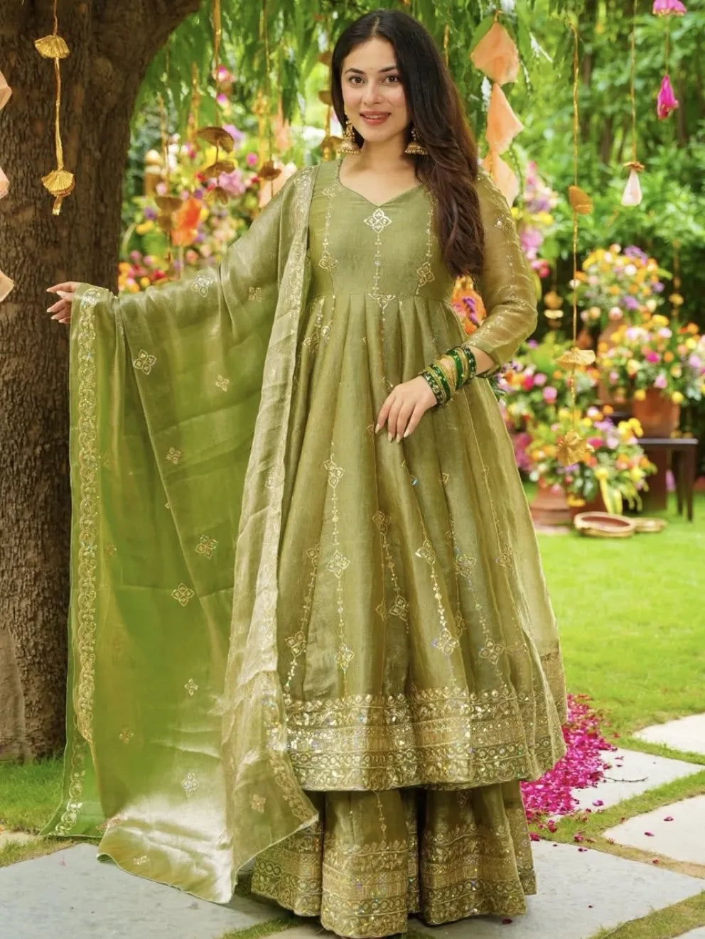 Woman in a green traditional outfit standing outdoors with flowers and decorations in the background