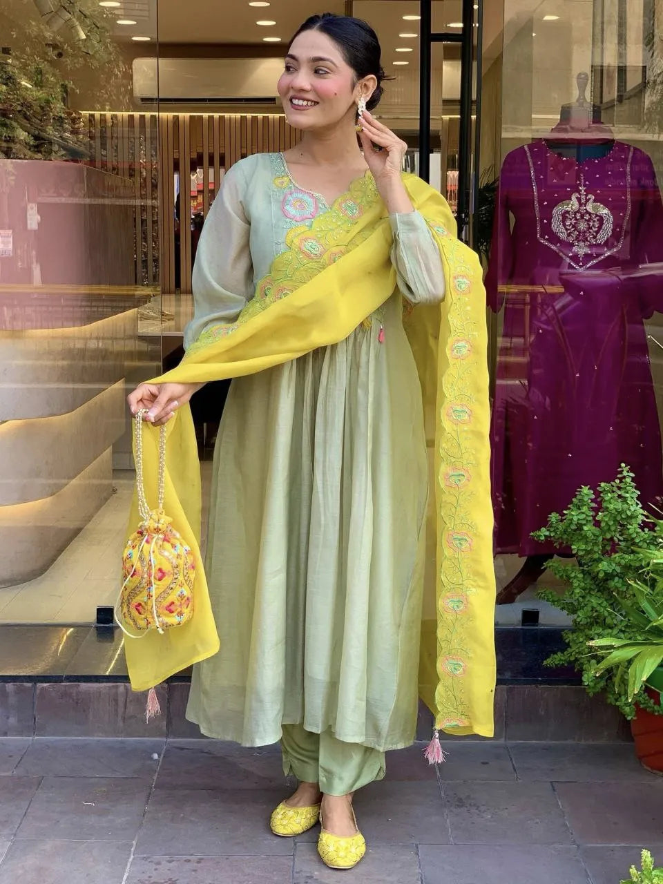 Woman in a light green traditional outfit with yellow dupatta standing in front of a store.